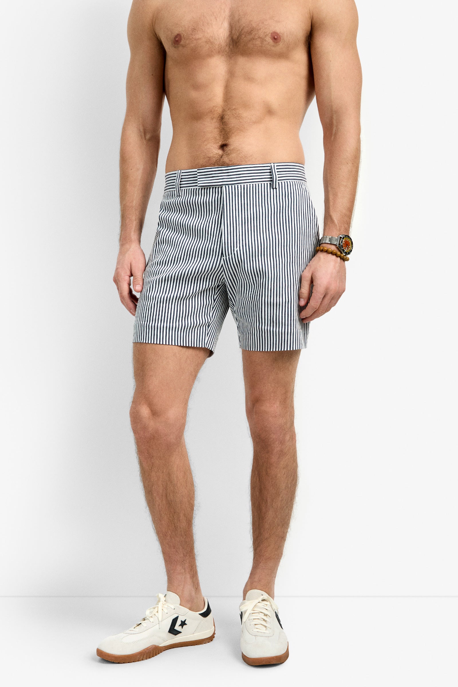 A shirtless man wears Roger Short in navy and white vertical stripes, paired with white sneakers featuring a navy logo and a wristwatch, standing against a plain white background.