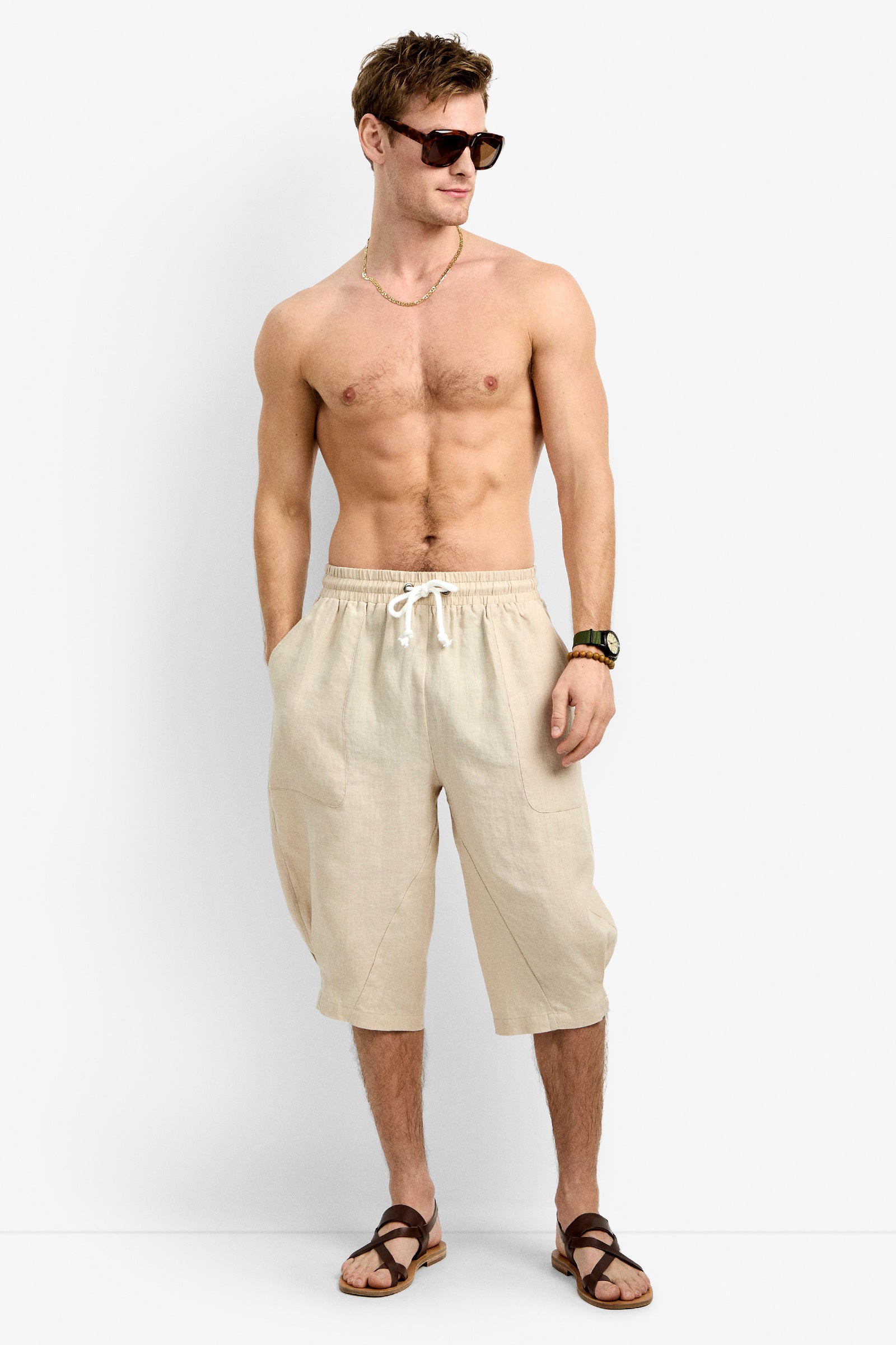 A shirtless man wears Claude Wide Leg Short in BEACHY BEIGE, brown sandals, sunglasses, a watch, and a necklace as he stands against a plain white background looking to the side with a relaxed posture.