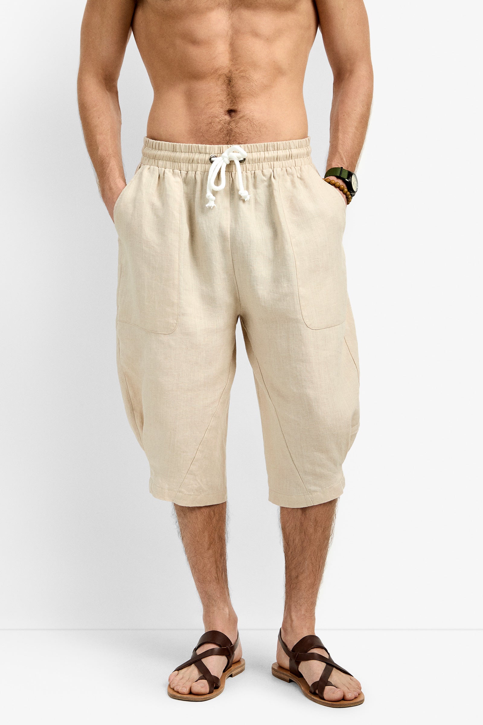 A man stands against a plain background, hands in his pockets, wearing the Claude Wide Leg Short—knee-length beige linen shorts with a white drawstring—paired with brown sandals and a wristwatch, no shirt.