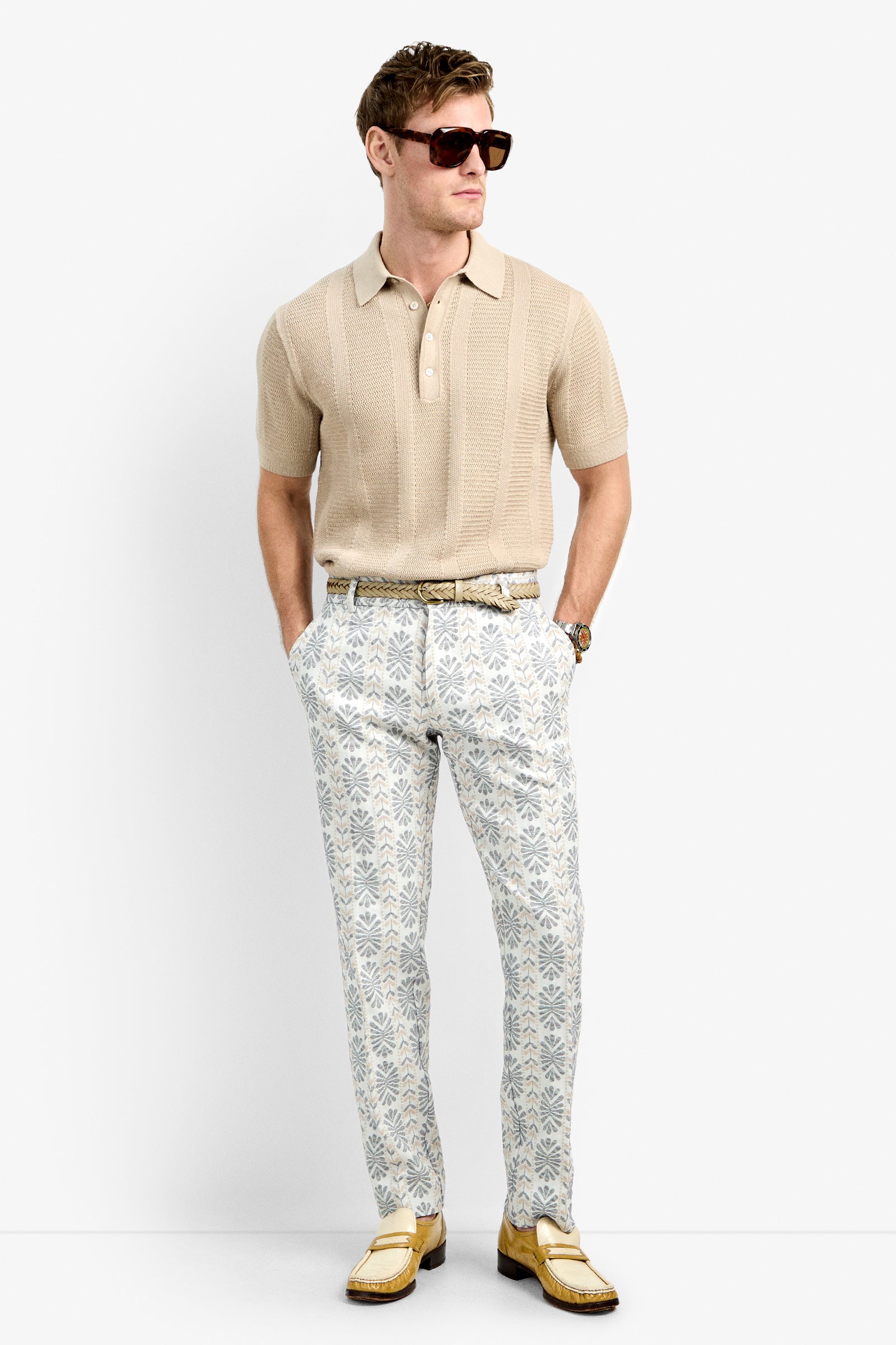 A man wearing a beige short-sleeve polo, Clyde Slim Trousers in patterned white and gray, yellow loafers, sunglasses, and a watch stands with his hands in his pockets against a plain white background.