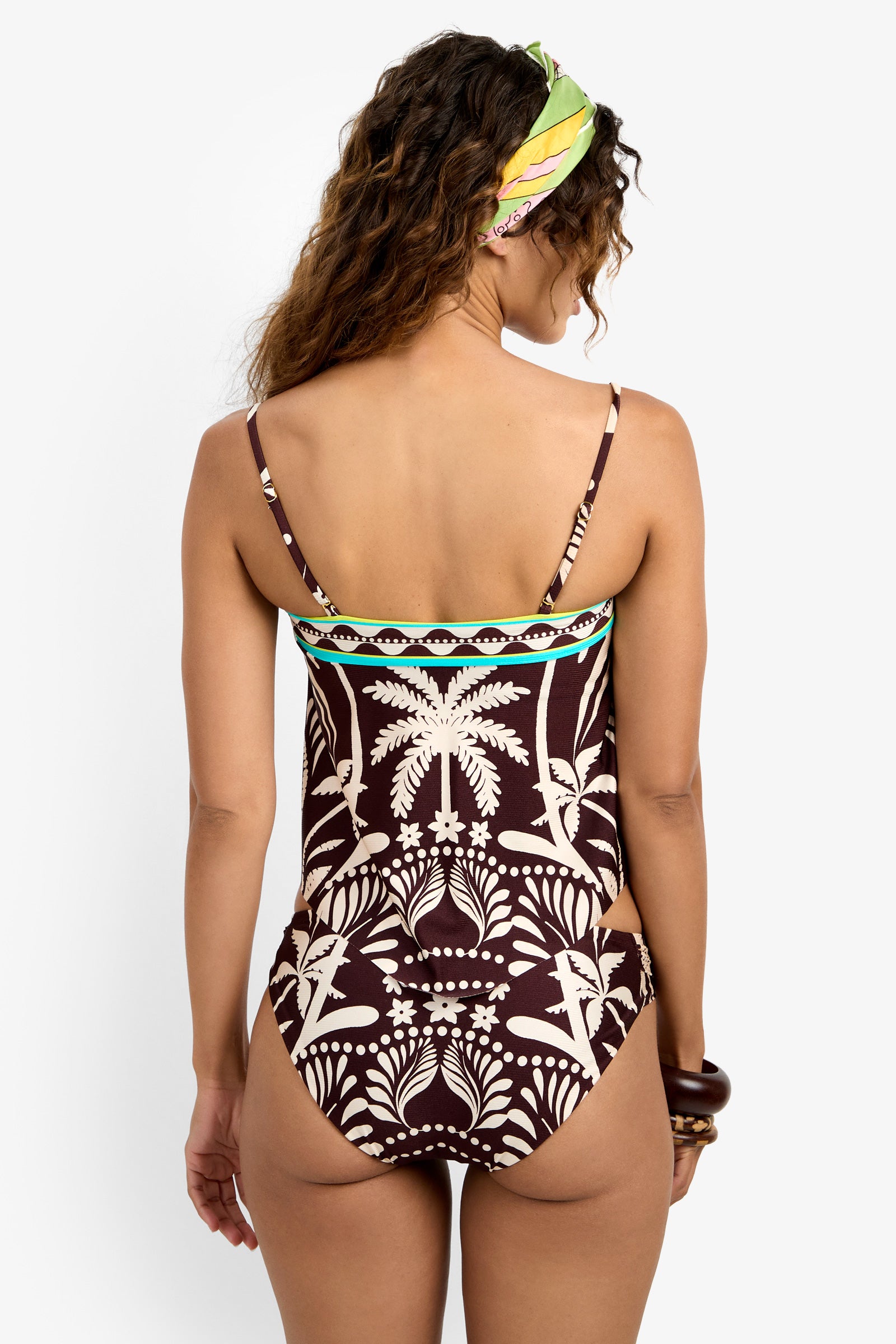 A woman with wavy hair and a colorful headscarf is seen from behind, wearing the Handkerchief Tankini Swim Top in brown with a white palm tree pattern and thin shoulder straps.