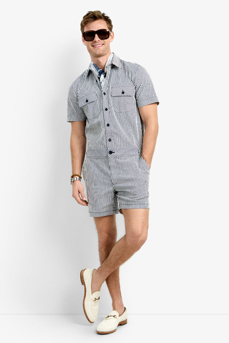 A man stands against a white background wearing sunglasses and the Sherman Short Jumpsuit— a short-sleeved, black-and-white striped button-up with matching shorts. He has one hand in his pocket and is wearing cream loafers.