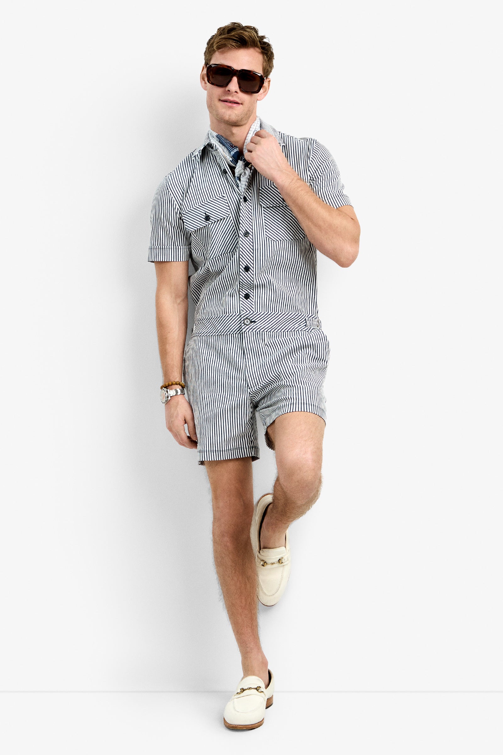 A man in the Sherman Short Jumpsuit, sunglasses, a watch, a blue patterned scarf, and cream loafers stands against a white background, leaning with one leg crossed.