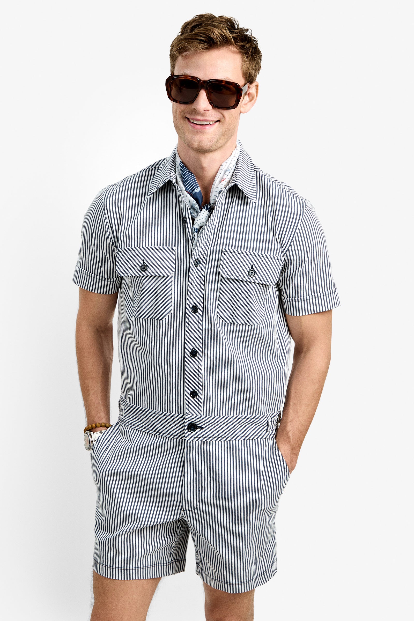 A man smiles with hands in his pockets, wearing sunglasses and the Sherman Short Jumpsuit—a blue and white striped short-sleeve one-piece—paired with a scarf, all against a plain white background.