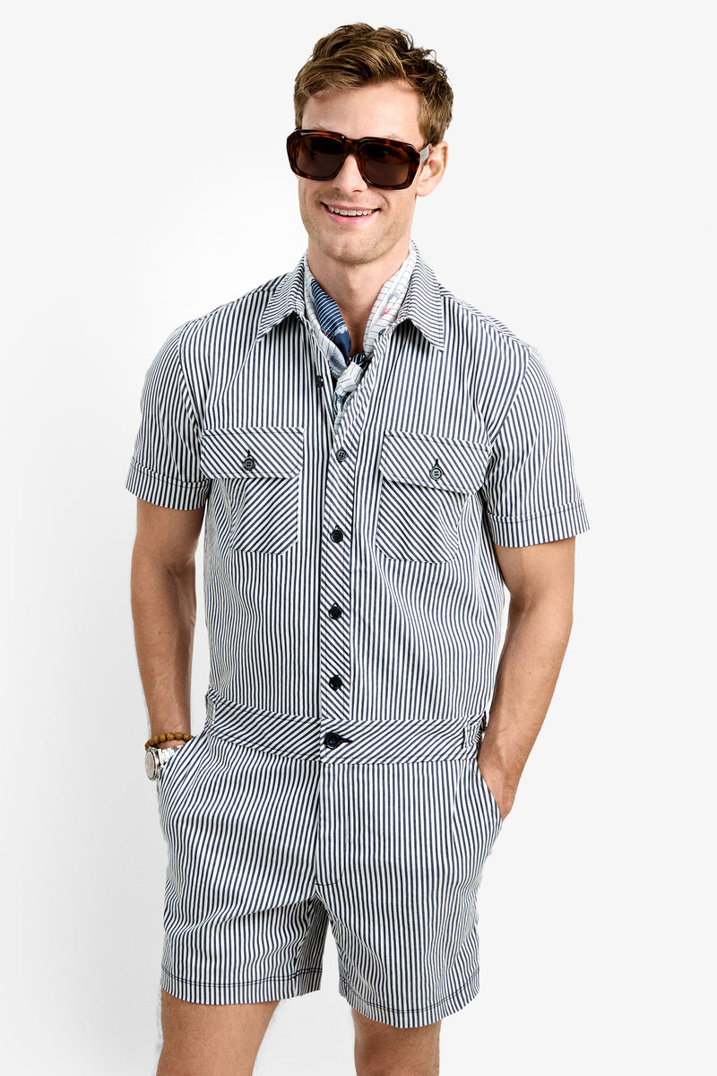 A man smiles with hands in his pockets, wearing sunglasses and the Sherman Short Jumpsuit—a blue and white striped short-sleeve one-piece—paired with a scarf, all against a plain white background.