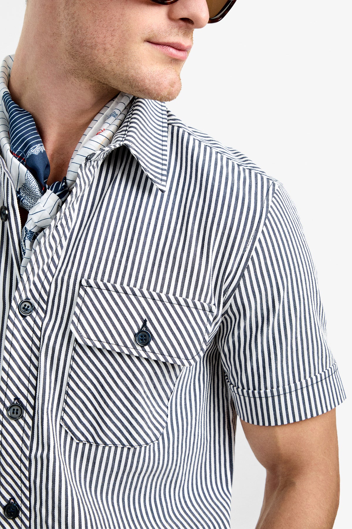 A person wearing sunglasses and the Sherman Short Jumpsuit with a blue and white striped pattern, short sleeves, front pocket, and a neck scarf, looks to the side against a plain background.