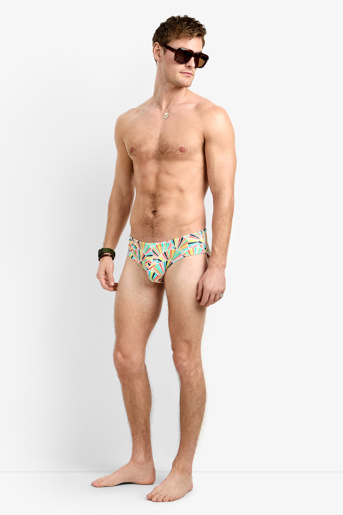 A man stands barefoot against a white background, wearing sunglasses, a necklace, and the Bondi Swim Brief—colorful and patterned. He looks to the side with a relaxed posture.