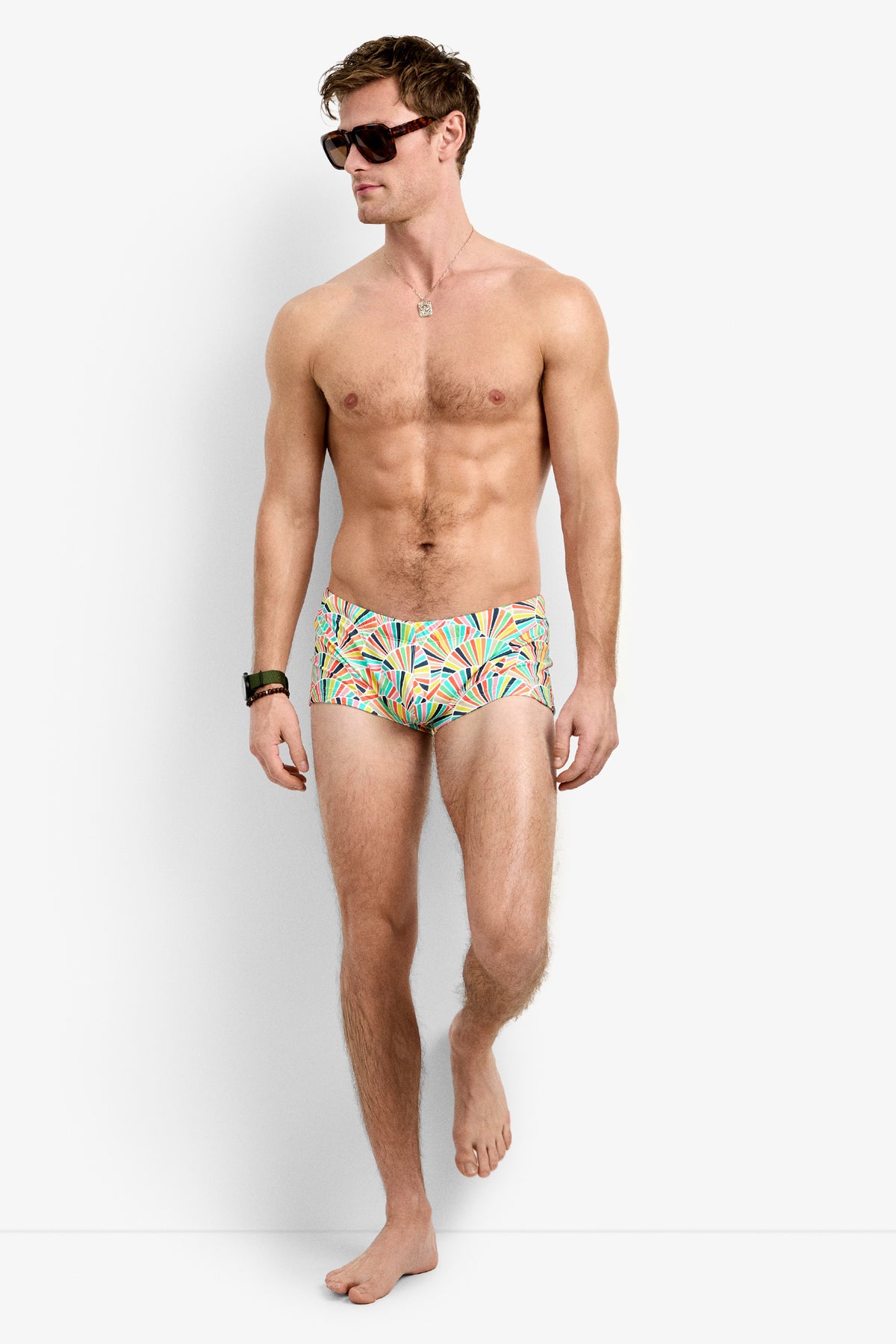 A shirtless man in Sorrento Swim Trunk and sunglasses stands on one foot against a plain white background, looking to his left.