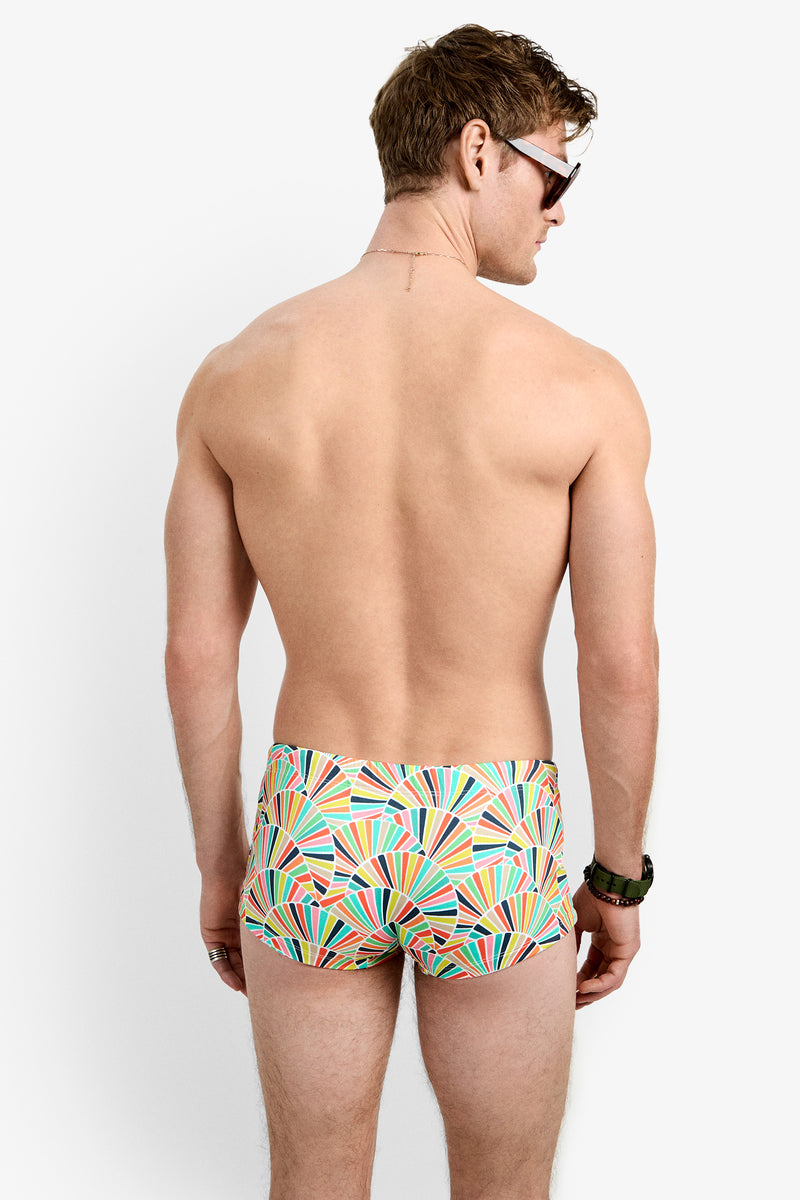 A man stands with his back to the camera, wearing Sorrento Swim Trunk featuring colorful geometric patterns, along with sunglasses and a watch, set against a plain white background.