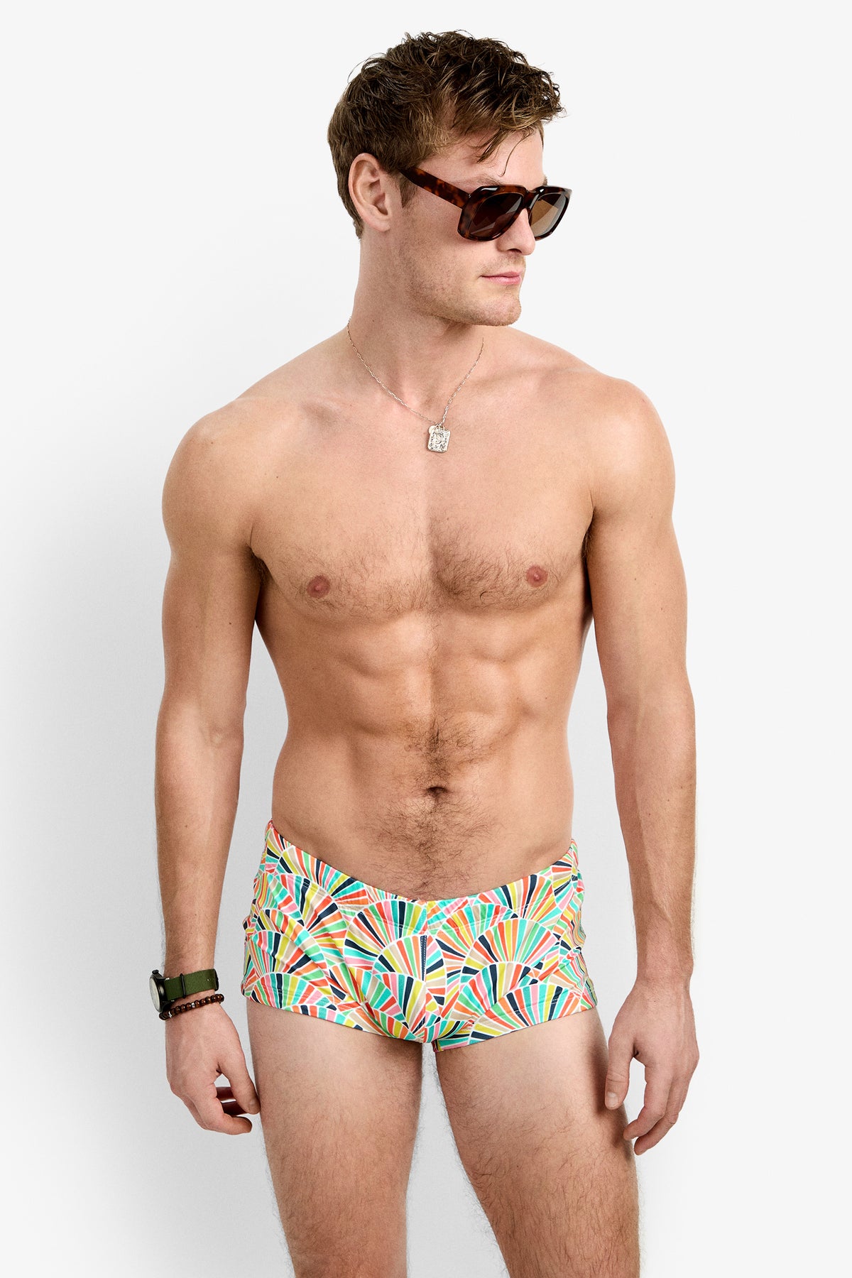 A man wearing Sorrento Swim Trunk with colorful geometric patterns, sunglasses, a wristwatch, and a necklace stands against a plain white background, looking to his right.