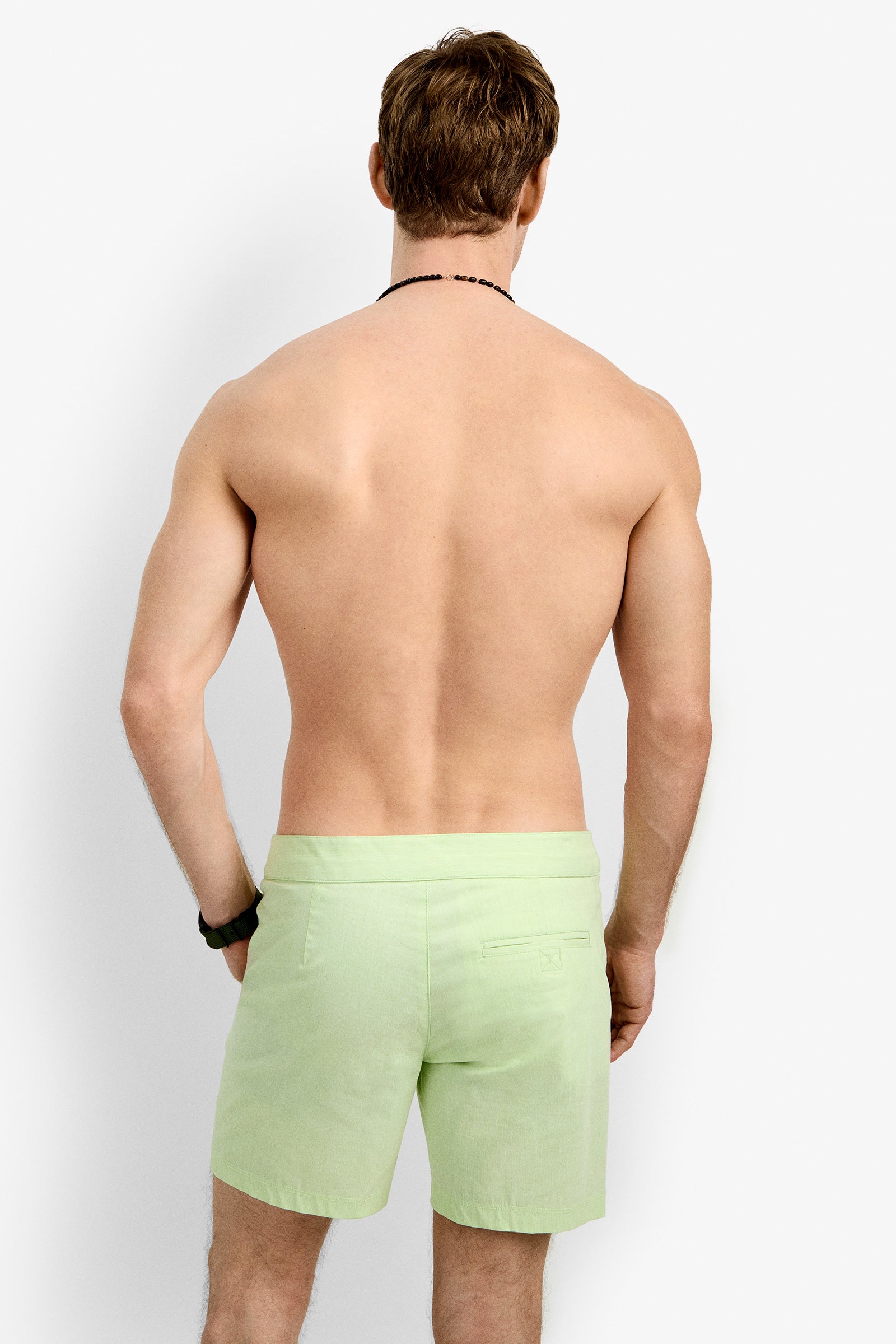 A shirtless, light-skinned man with brown hair stands facing away in Honolulu Board Short (light green) and a black wristband, set against a plain white background.