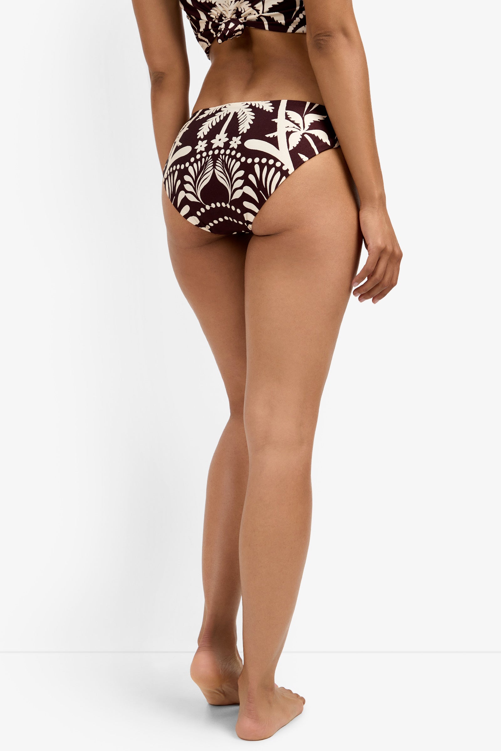A woman stands barefoot on a white background, modeling the Tab Side Hipster Swim Bottom in a dark brown and cream tropical pattern, shown from behind with her right arm relaxed at her side.