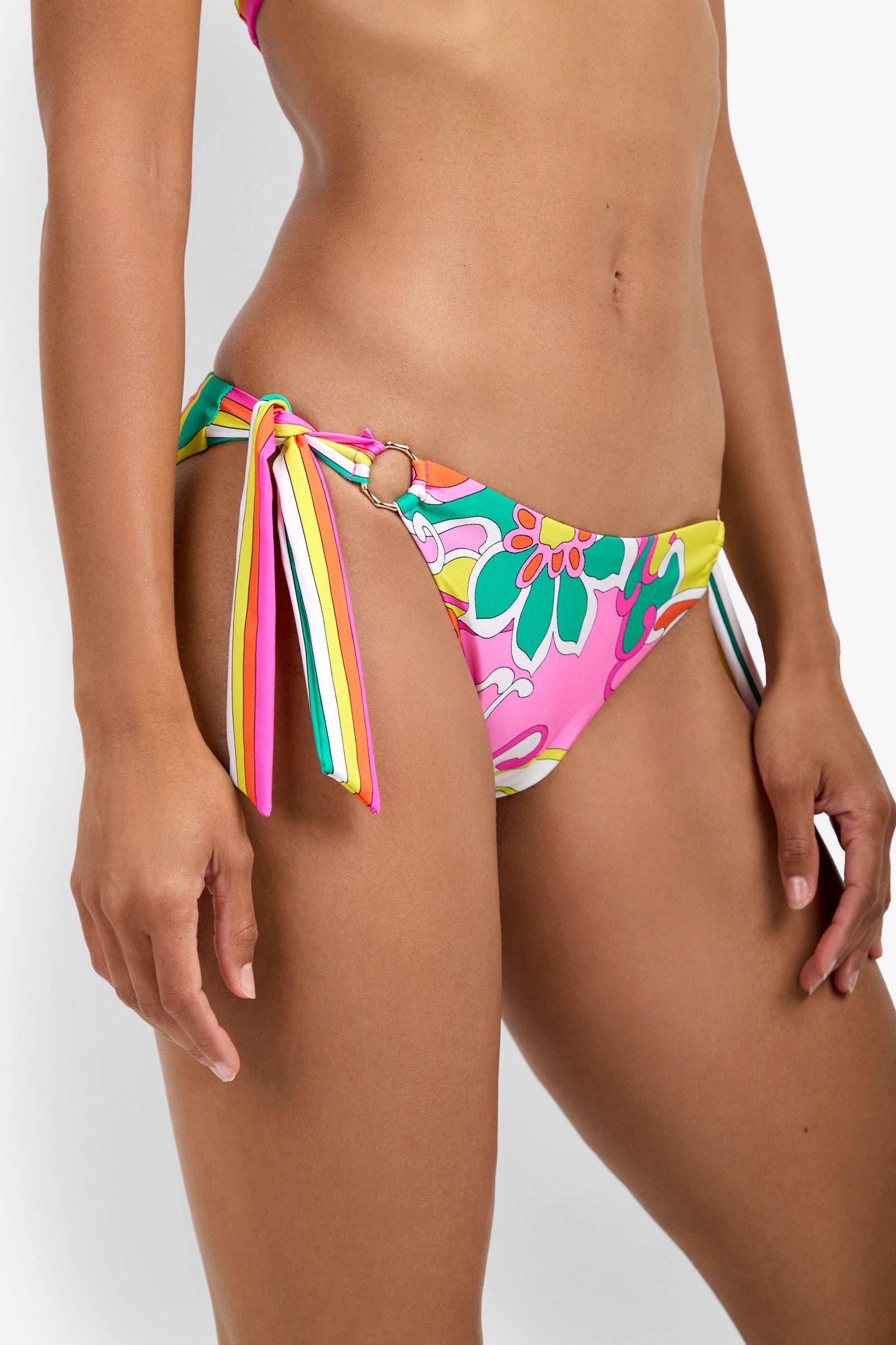 A woman models the Ring Hipster Swim Bottom, featuring a colorful floral print in pink, green, yellow, and white with side ties and a ring detail at the hip. Only her torso and upper thigh are visible.