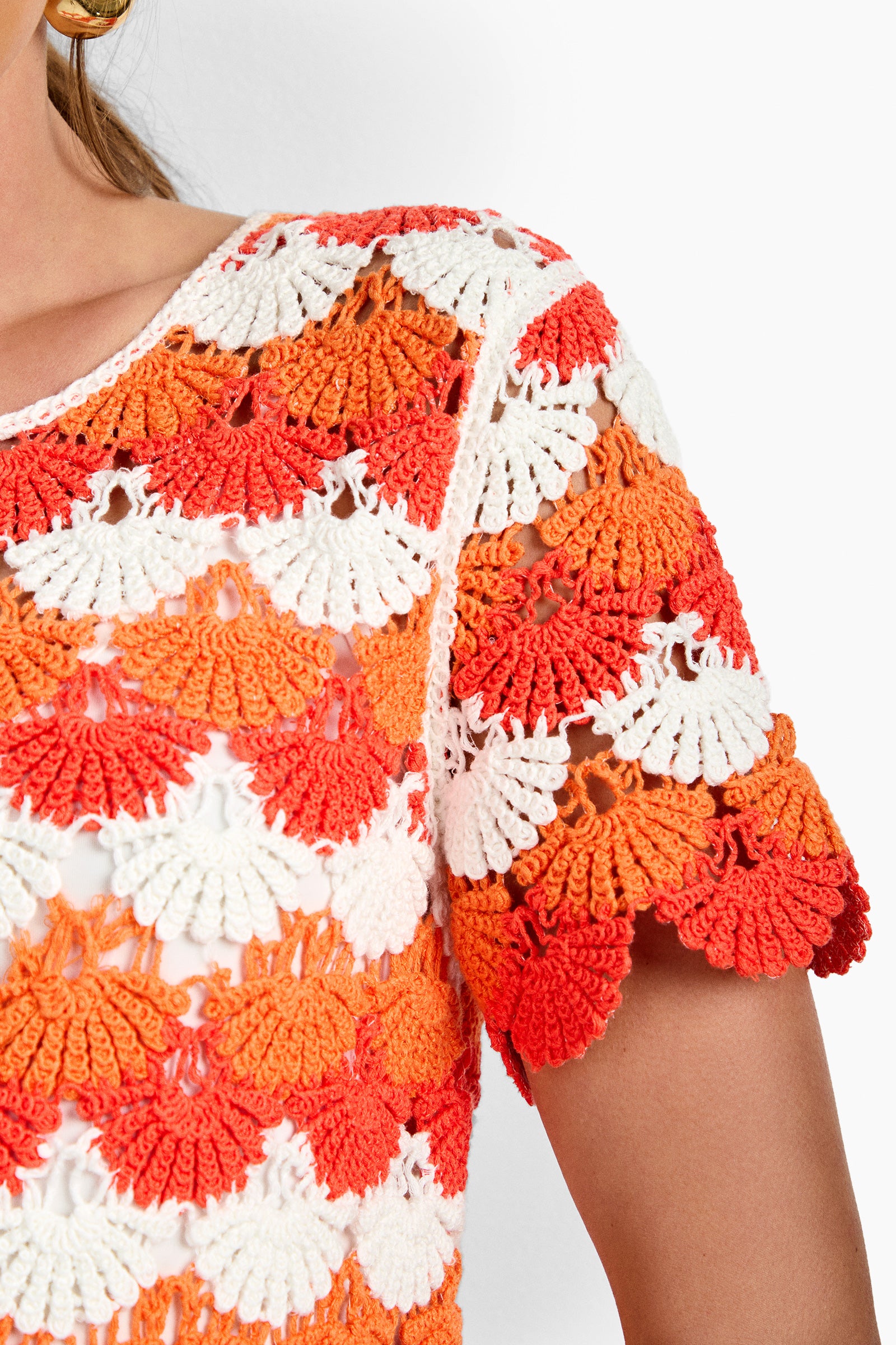Close-up of a woman's shoulder wearing the Solana Beach Dress—a crocheted top with scalloped orange, white, and red patterns, textured fan-like designs, and short sleeves.