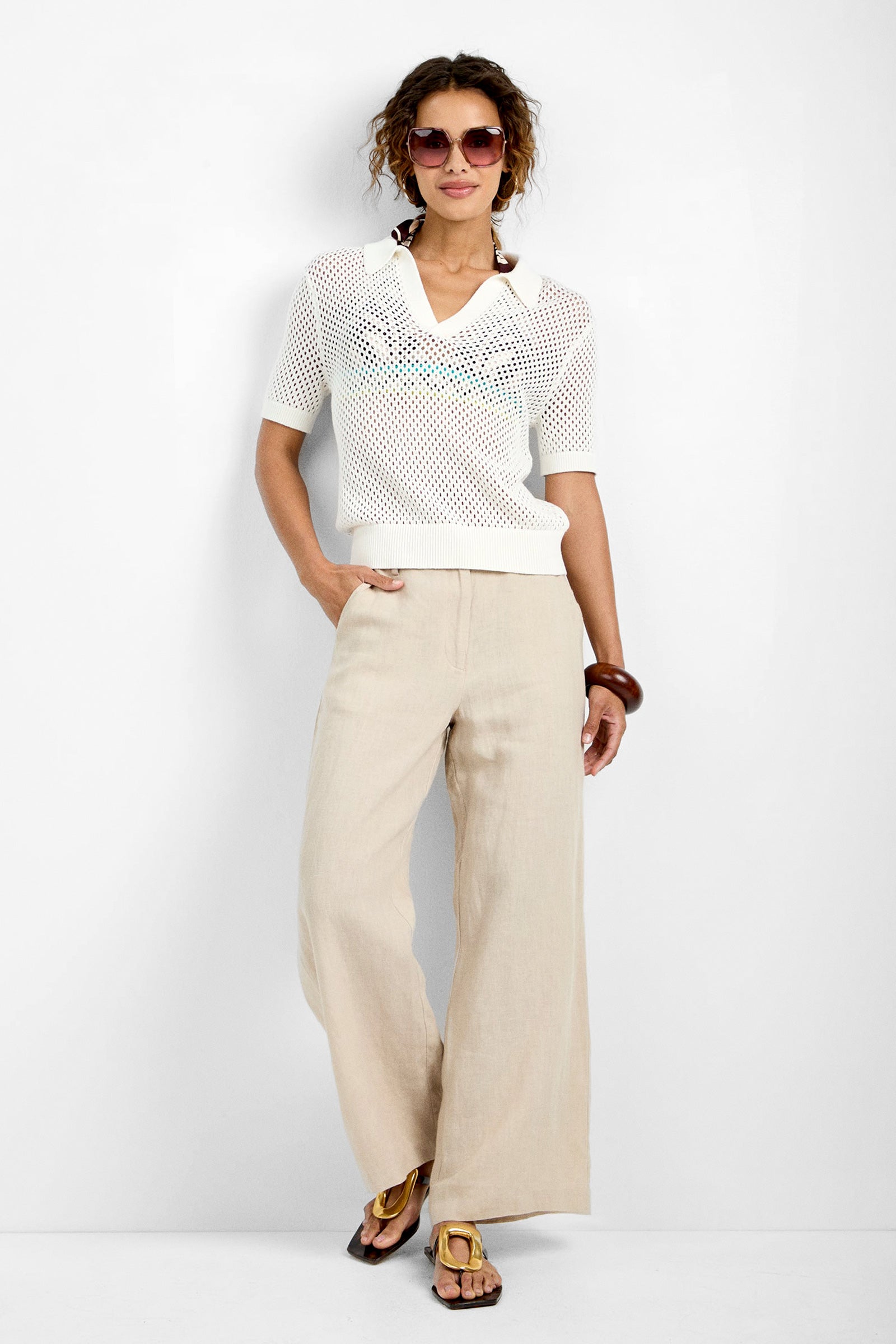 A woman stands against a white background wearing TEST-1 with sunglasses, beige wide-leg linen pants, sandals, and a brown bangle, one hand in her pocket.