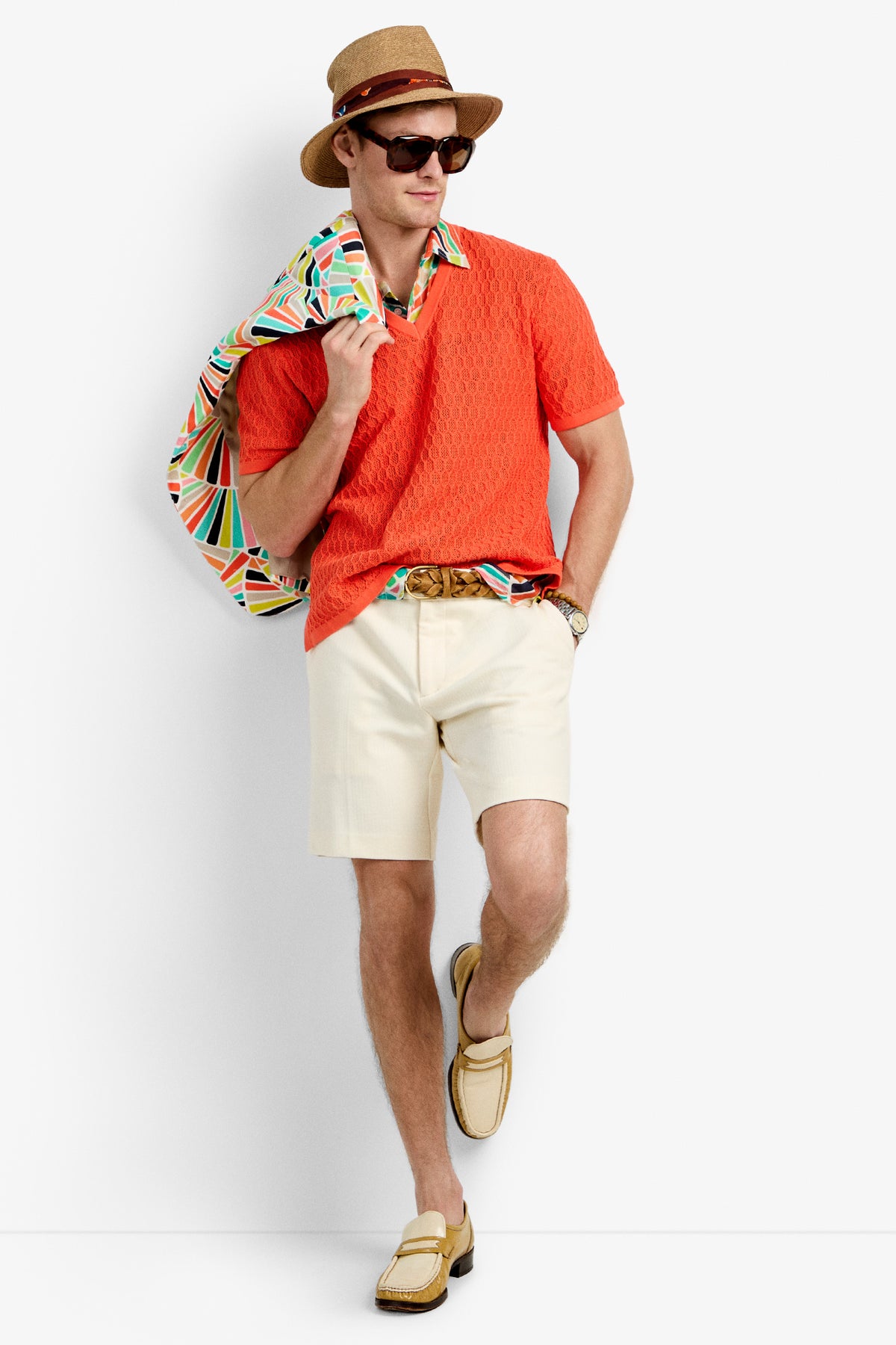 A man poses stylishly in dark sunglasses, a straw hat, cream shorts, beige loafers, a patterned belt, and holds a colorful jacket over his shoulder while wearing the Heyes Mesh Henley V-Neck in bright orange.