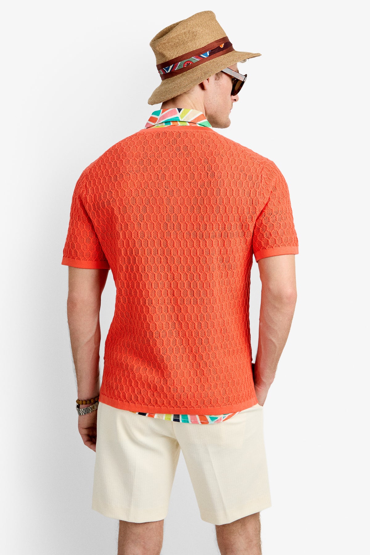 A man faces away, wearing the Heyes Mesh Henley V-Neck in orange, layered over a colorful shirt collar. He pairs it with light shorts, a tan hat with a patterned band, sunglasses, and bracelets.