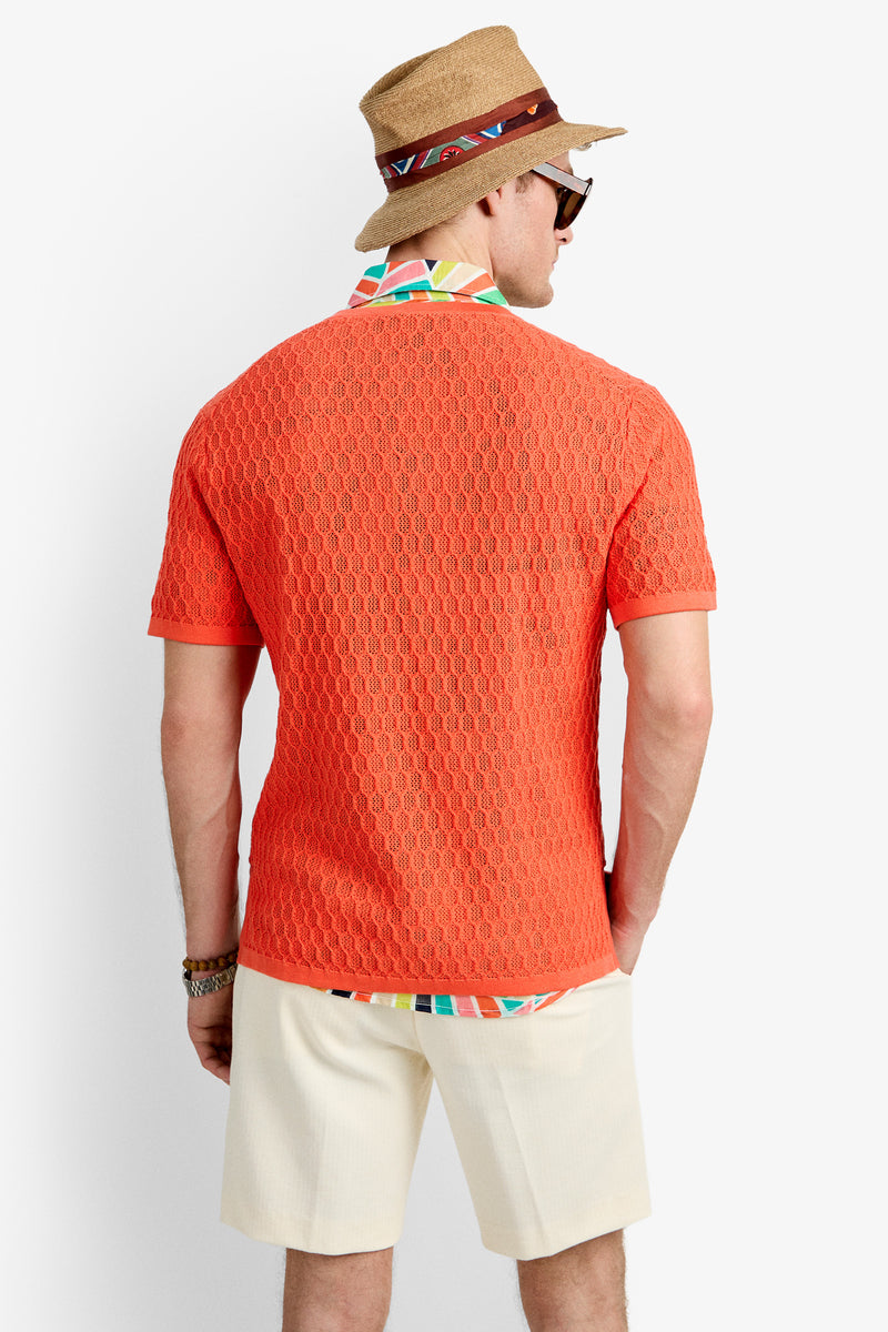 A man faces away, wearing the Heyes Mesh Henley V-Neck in orange, layered over a colorful shirt collar. He pairs it with light shorts, a tan hat with a patterned band, sunglasses, and bracelets.