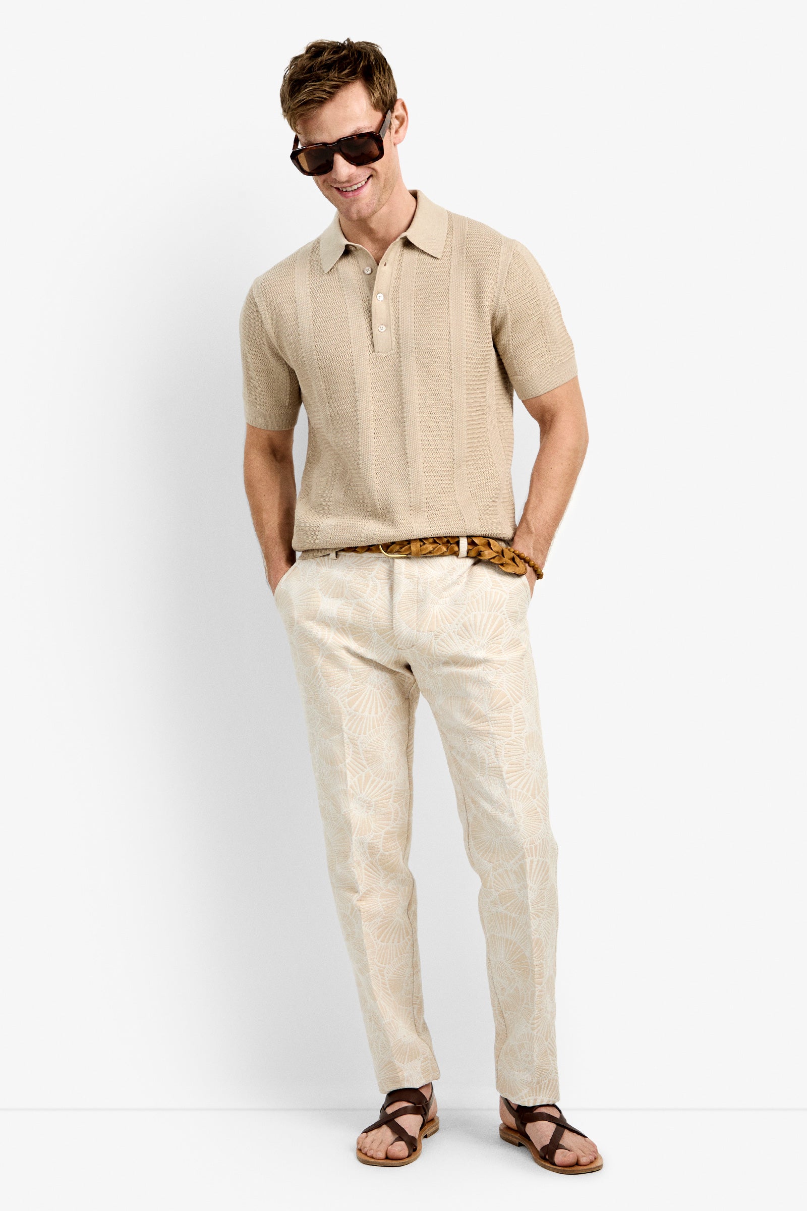 A man wears the Devan Short Sleeve Polo in beige with light patterned pants, a woven belt, brown leather sandals, and dark sunglasses, smiling with his hands in his pockets against a plain white background.