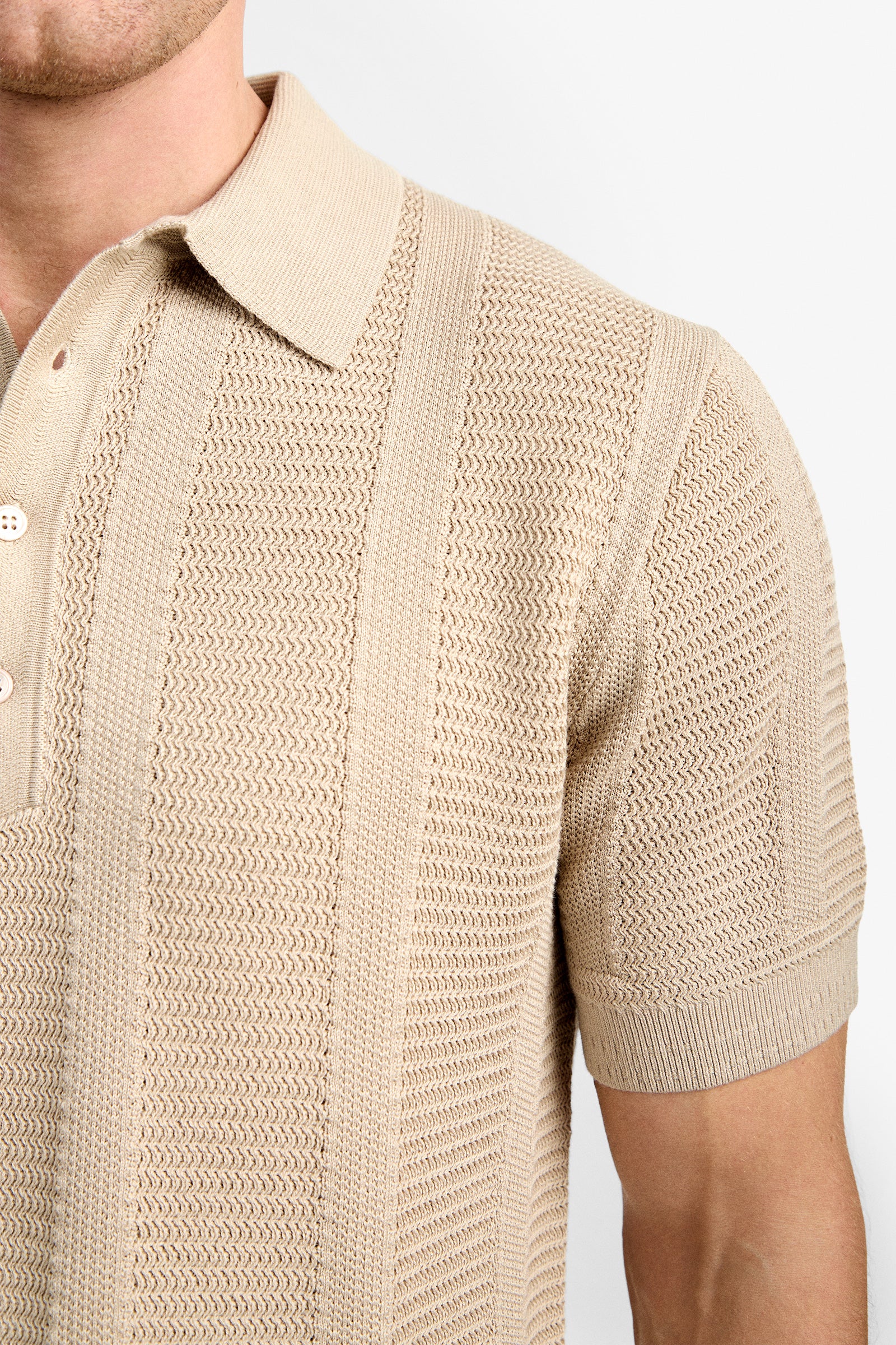 A close-up of a person wearing the Devan Short Sleeve Polo, a beige knit shirt with a collar and buttons, highlighting the textured pattern of the fabric.