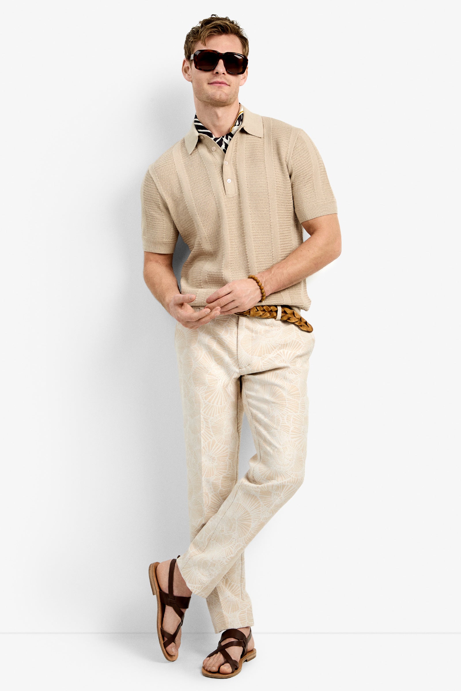 A man wearing the Devan Short Sleeve Polo in beige, patterned cream trousers, brown sandals, sunglasses, and a striped neck scarf stands against a white background with his arms loosely crossed.