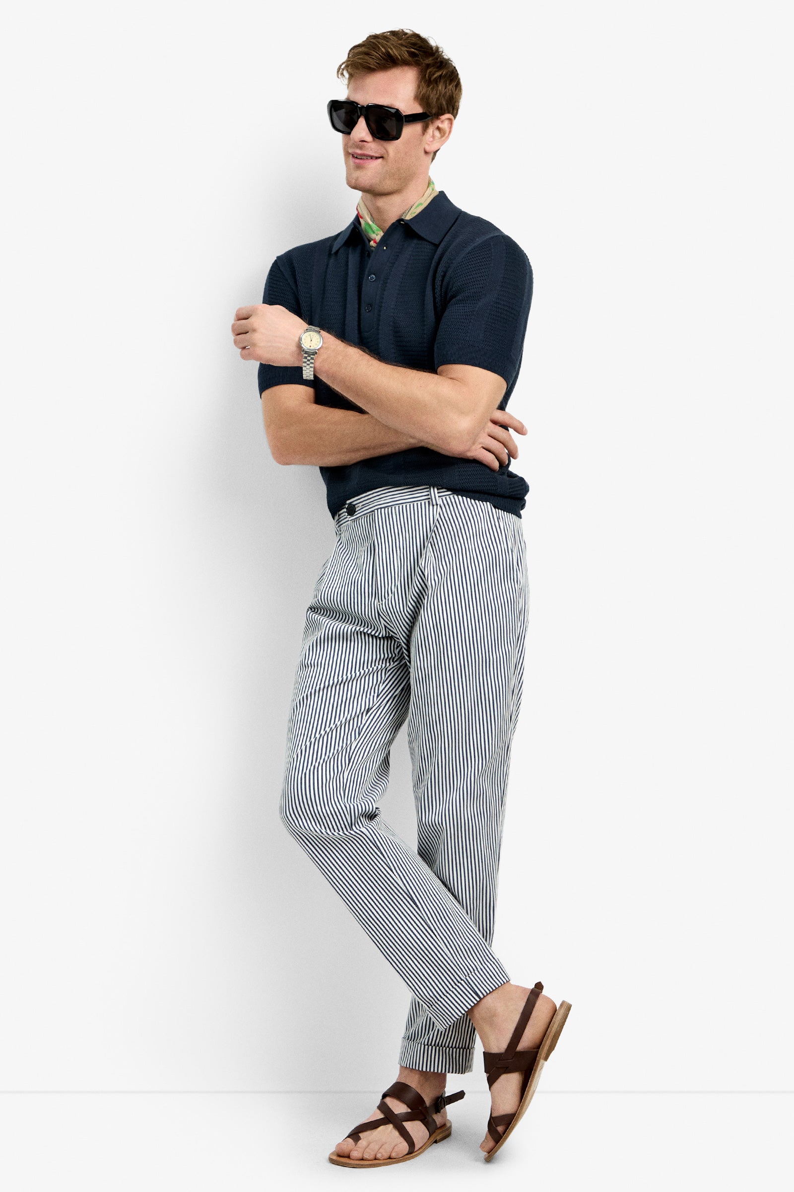 A man wearing the Devan Short Sleeve Polo stands against a white background with sunglasses, striped pants, brown sandals, arms crossed, and legs casually crossed.