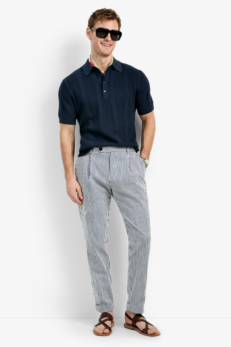 A man smiles with one hand in his pocket, wearing the Devan Short Sleeve Polo in navy, dark sunglasses, light striped pants, and brown sandals against a plain white background.