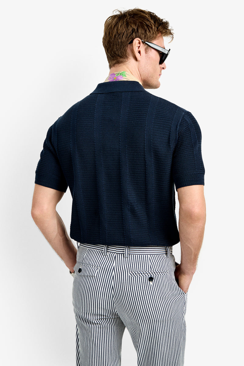 A man with short brown hair and sunglasses is seen from the back, wearing the Devan Short Sleeve Polo and high-waisted, black-and-white striped pants. He has a colorful neck tattoo and stands against a plain white background.