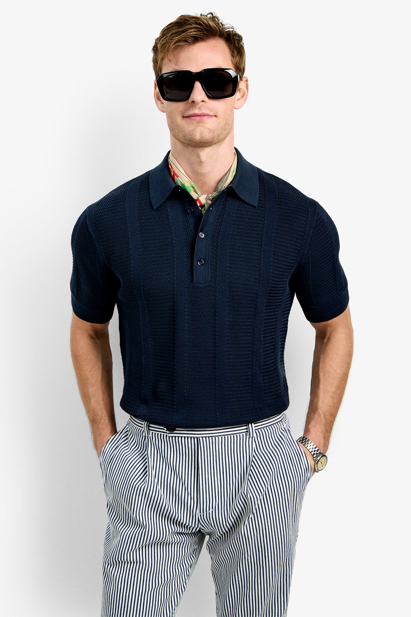 A man wearing the Devan Short Sleeve Polo, black sunglasses, a colorful neck scarf, and high-waisted striped pants stands with hands in his pockets against a plain white background.