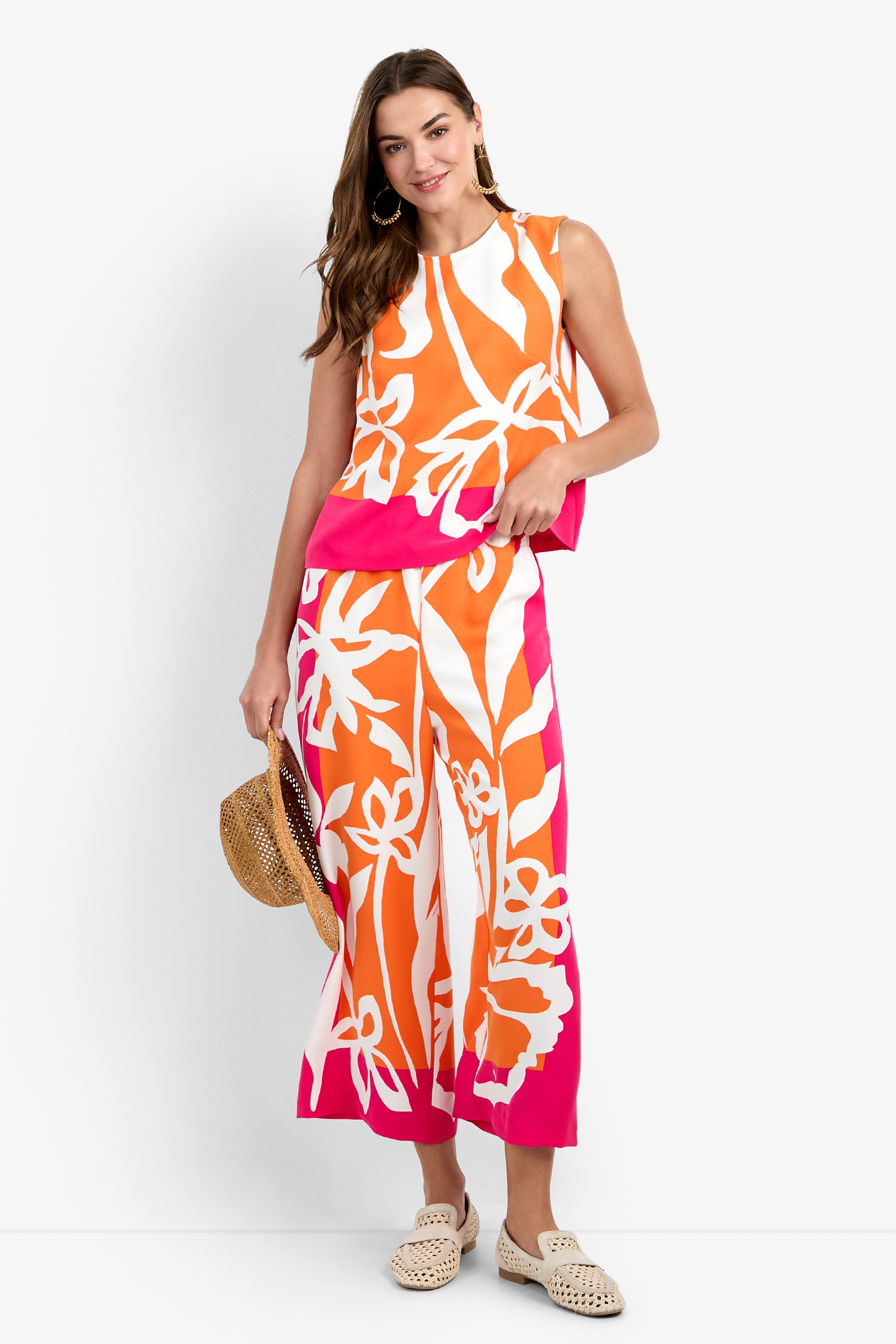 A woman wearing the Riggs Top, a sleeveless piece with a bold orange, pink, and white floral print, stands smiling and holding a straw hat. She pairs it with wide-leg pants, hoop earrings, long hair, and woven shoes.