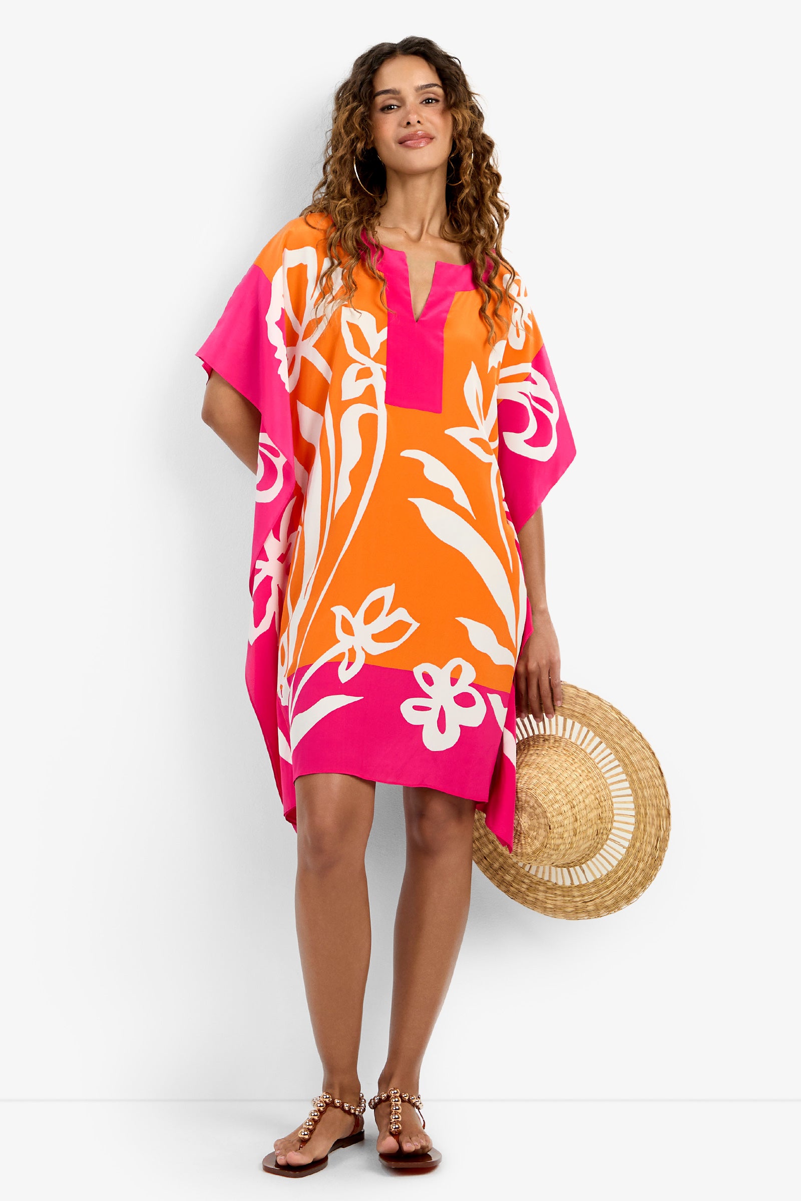 A woman with curly hair and a relaxed expression wears the Theodora Caftan Dress in orange, pink, and white floral print, holding a woven straw hat and standing in sandals.