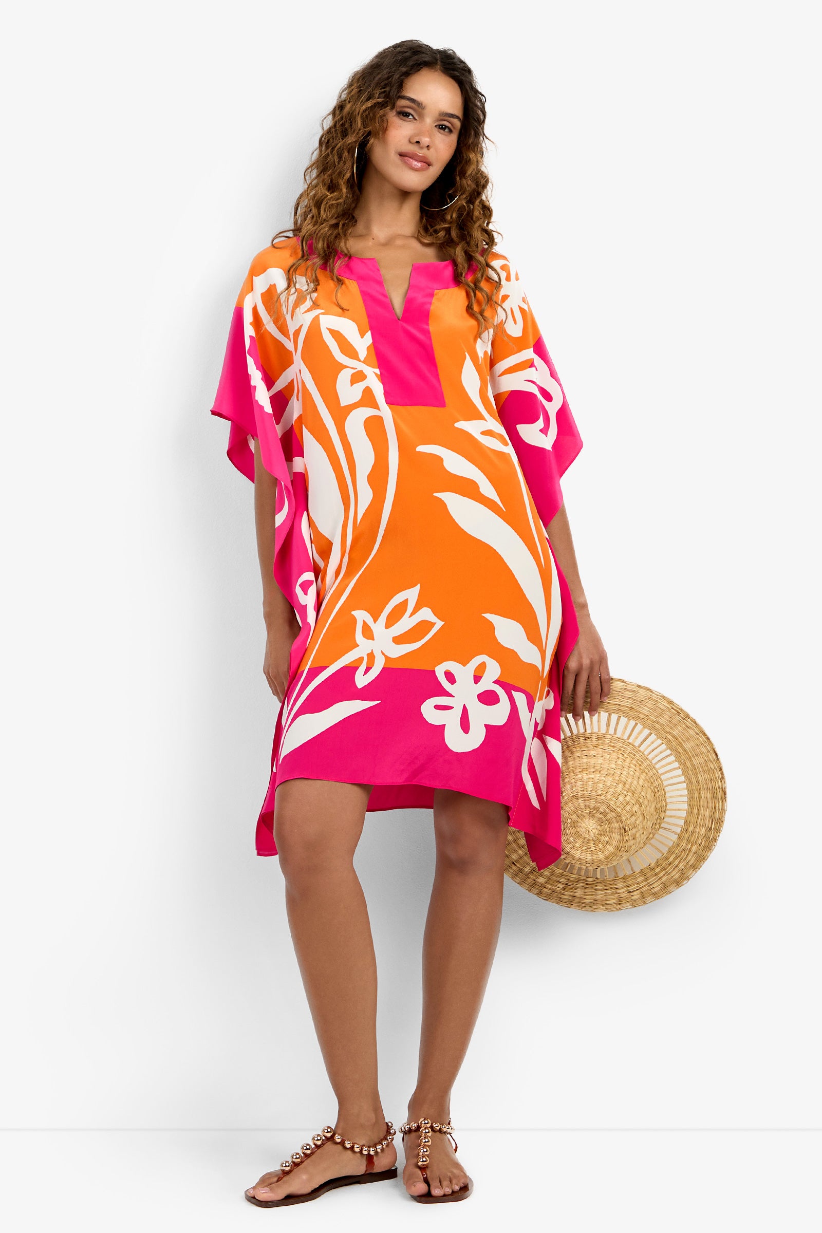 A woman with curly hair stands barefoot in a relaxed pose, wearing the Theodora Caftan Dress—an orange, pink, and white floral, loose-fitting style—paired with sandals and holding a round straw bag.