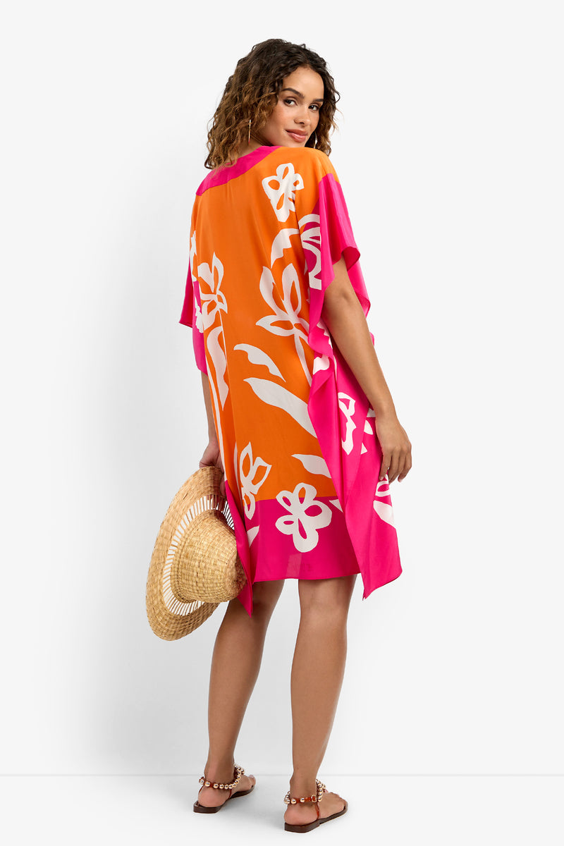 A woman faces away in the Theodora Caftan Dress, featuring loose, colorful orange, pink, and white florals. She holds a wide-brimmed straw hat, wears sandals, and her curly hair falls to her shoulders.
