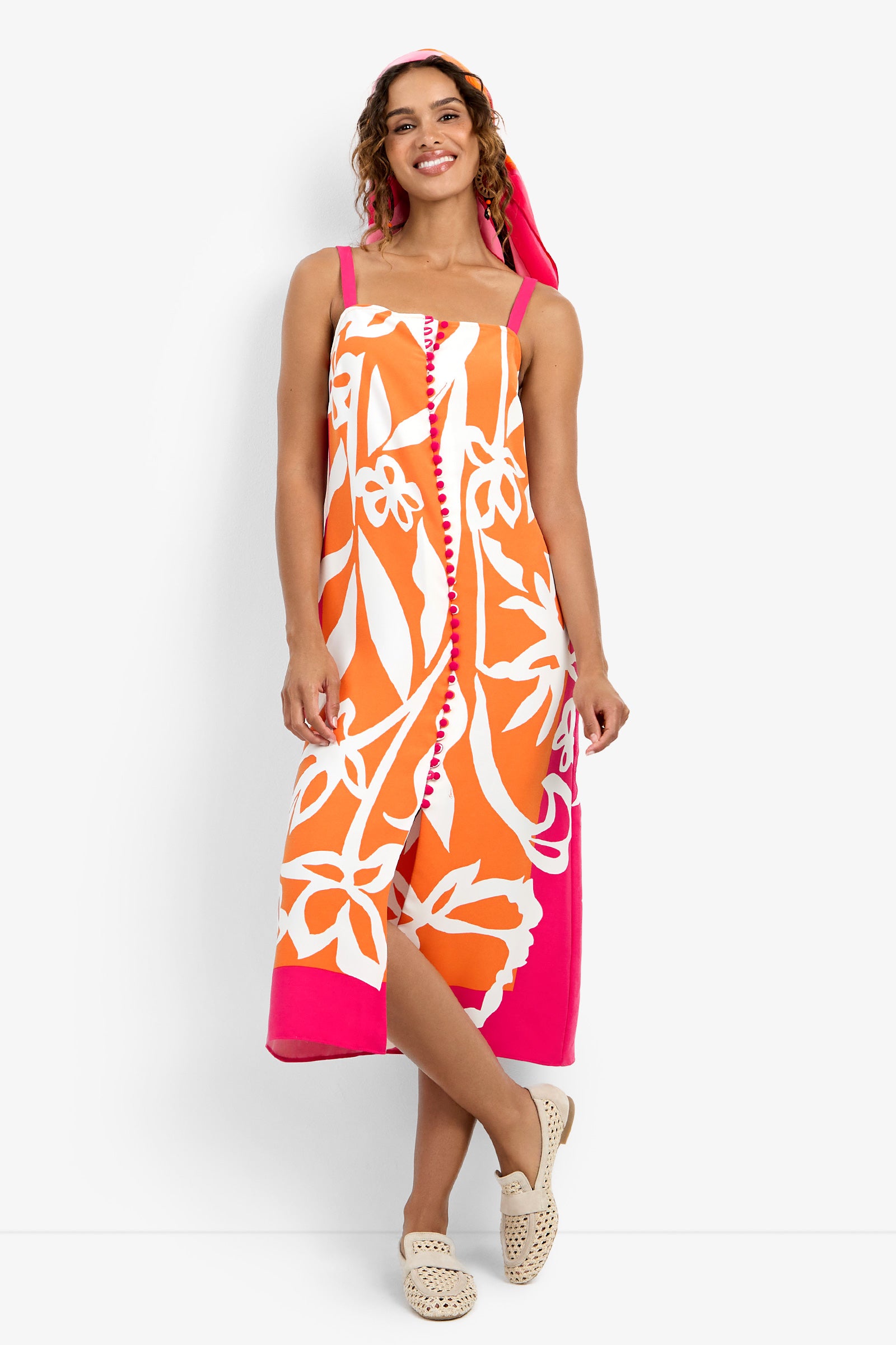 A woman smiles in the Haisley Dress, a sleeveless, mid-length style with bold orange, white, and pink floral patterns, paired with beige slip-on shoes and a matching pink headscarf.