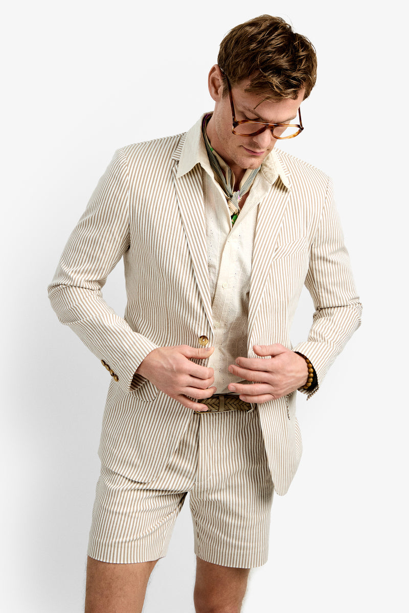 A man wearing glasses, the LOUIE BLAZER in beige and white stripes, matching shorts, a light shirt, and a patterned scarf around his neck looks down as he buttons his jacket.