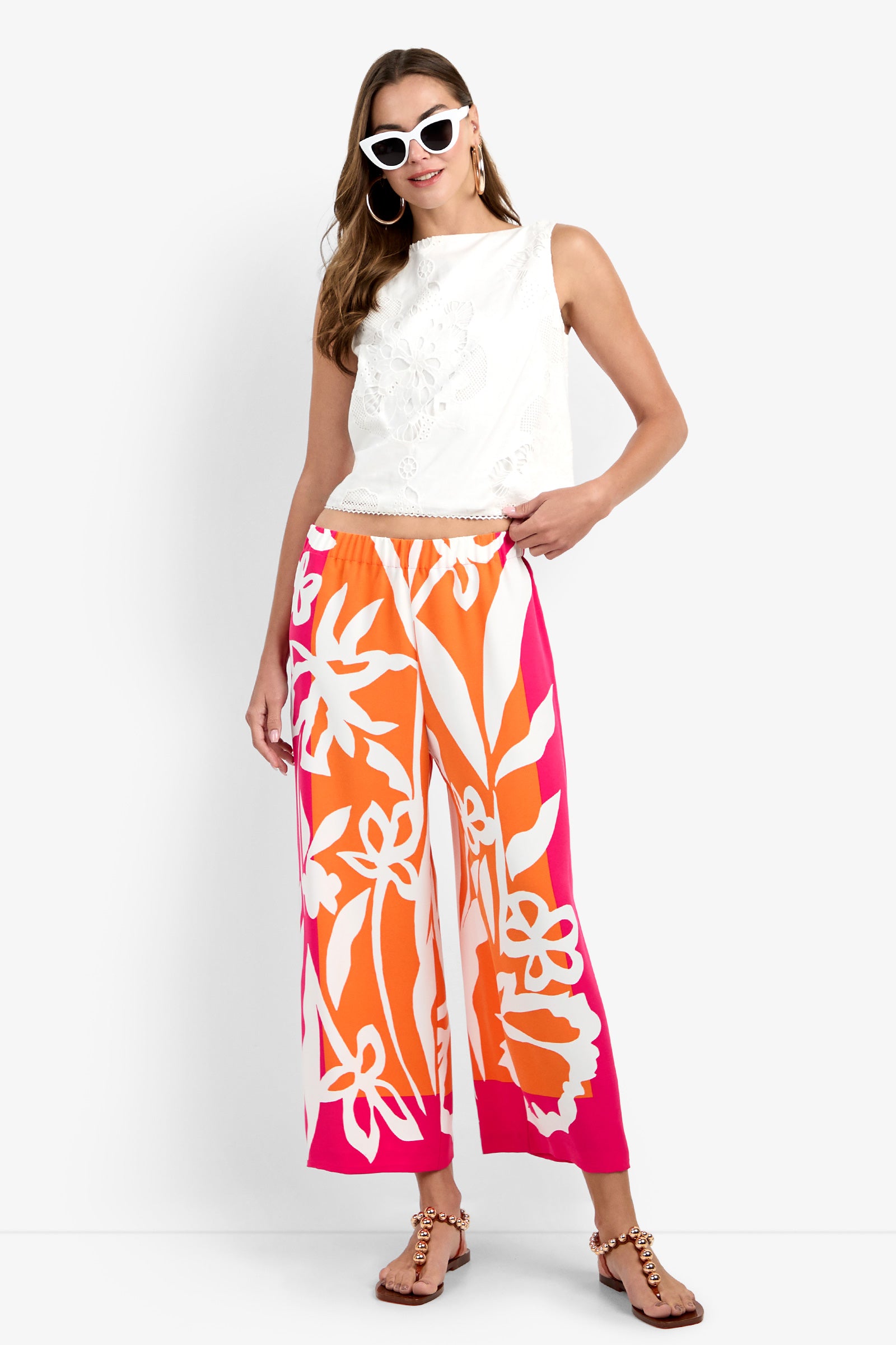 Woman wearing the Estella Pant—bold orange and pink floral wide-leg pants—with a white sleeveless top, cat-eye sunglasses, hoop earrings, and brown sandals, standing against a plain white background.
