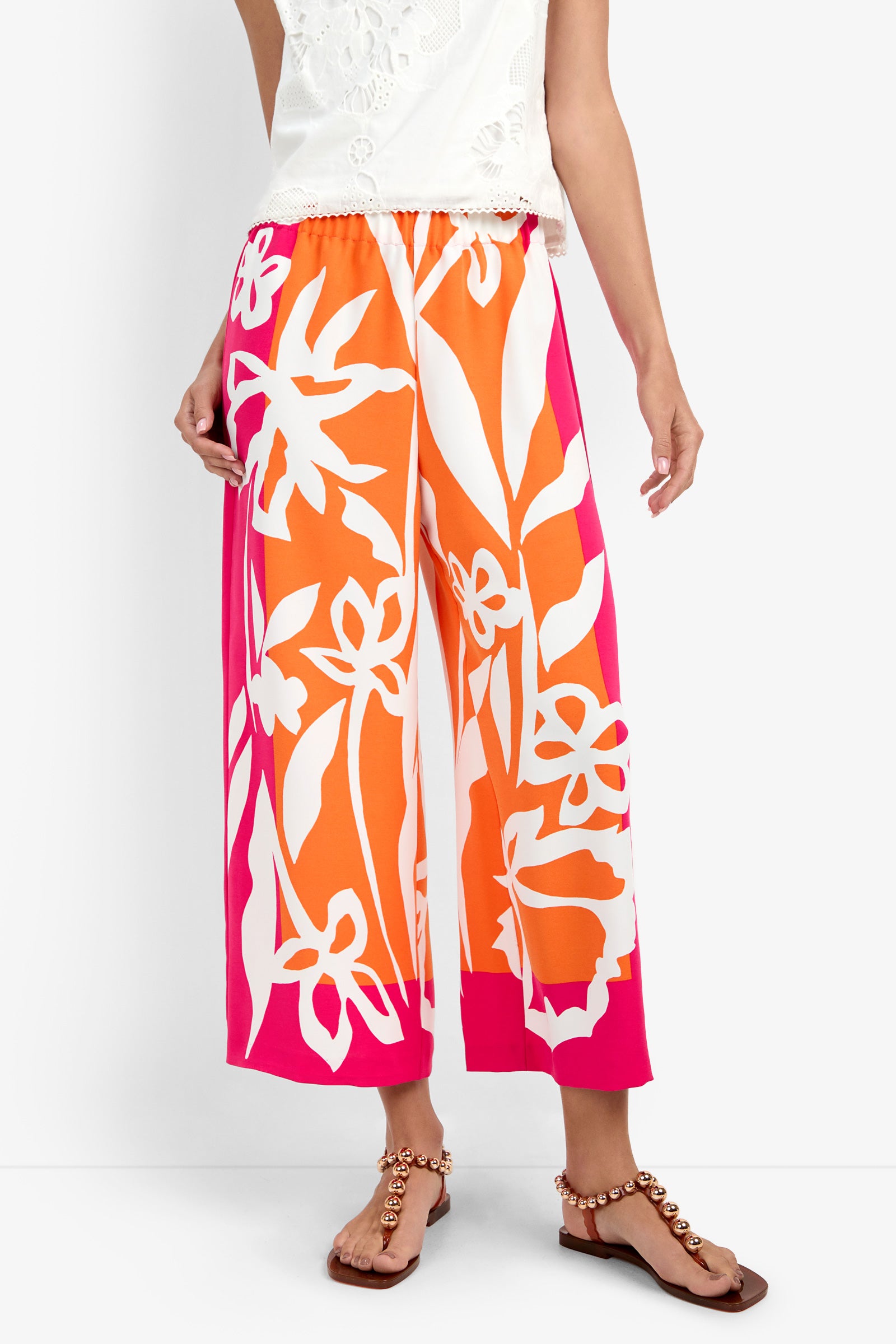 A person wearing the Estella Pant, featuring a wide-leg, cropped silhouette with bold orange, pink, and white abstract florals, styled with a white top and flat beaded-strap sandals.