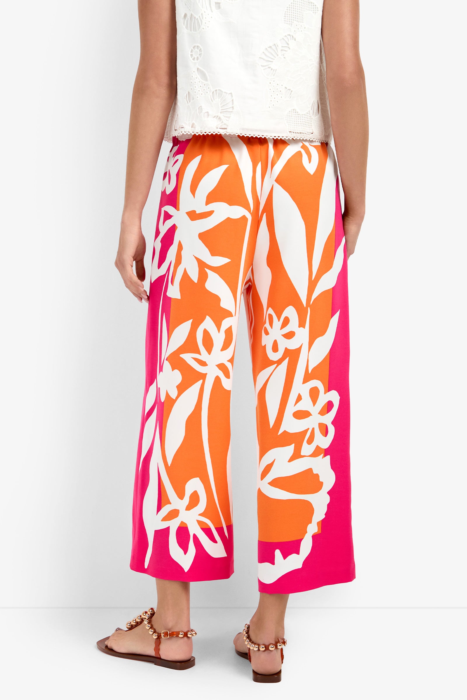 A person seen from the back wears the Estella Pant—wide-leg, ankle-length with a vibrant orange and pink floral print—paired with a white textured sleeveless top and flat sandals, set against a plain backdrop.