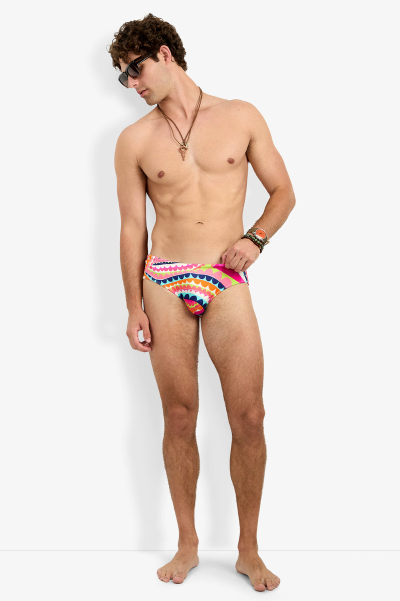 A shirtless man wearing sunglasses and the Bondi Swim Brief, featuring a colorful pattern, stands barefoot against a white background looking to his left, accessorized with necklaces and bracelets.