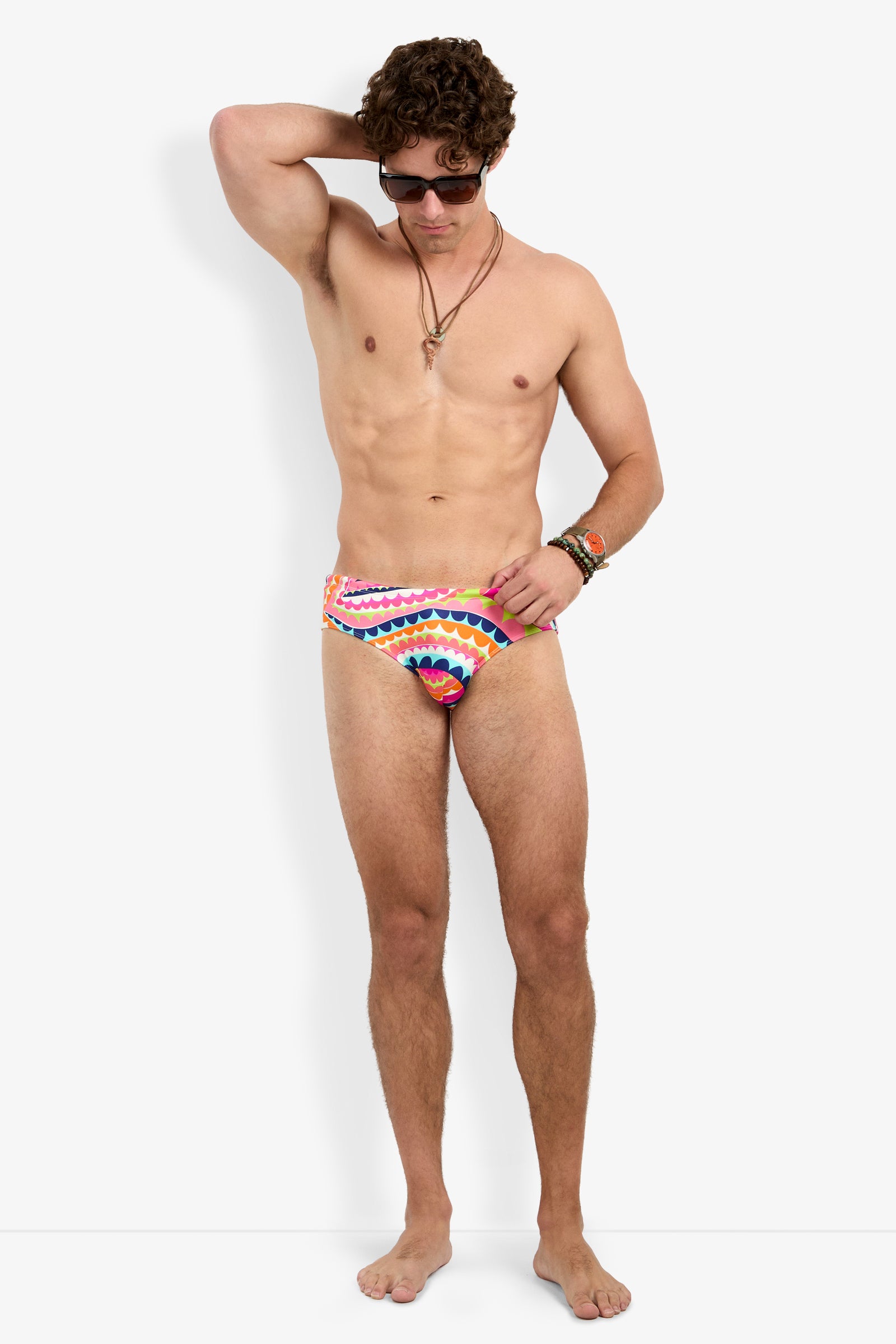 A man stands barefoot against a white background, wearing sunglasses, necklaces, and the Bondi Swim Brief in a colorful pattern. One hand is behind his head and the other rests by his side.