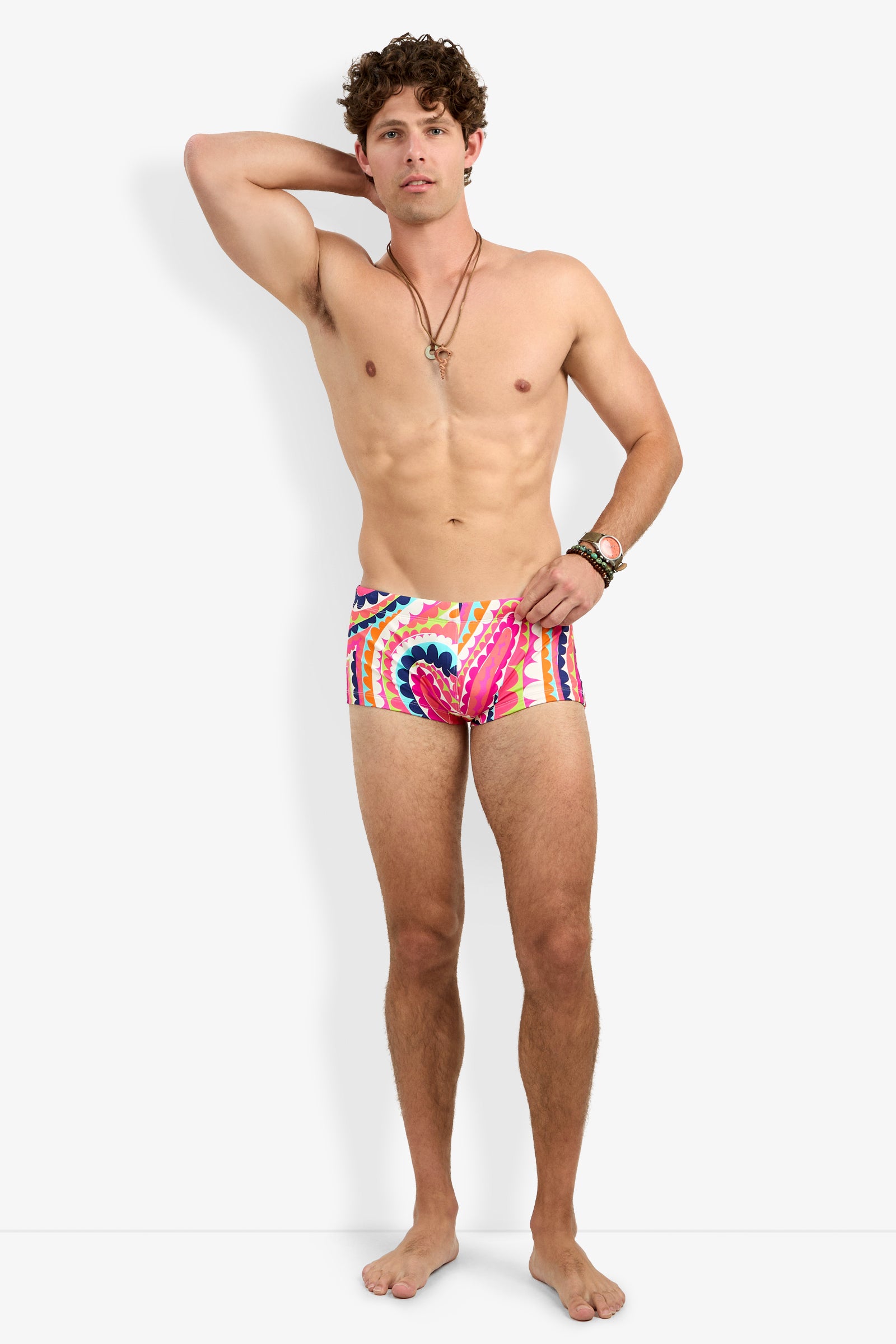 A shirtless man with curly hair stands barefoot against a white background, wearing Sorrento Swim Trunk. He accessorizes with necklaces and bracelets, one hand behind his head and the other by his side.