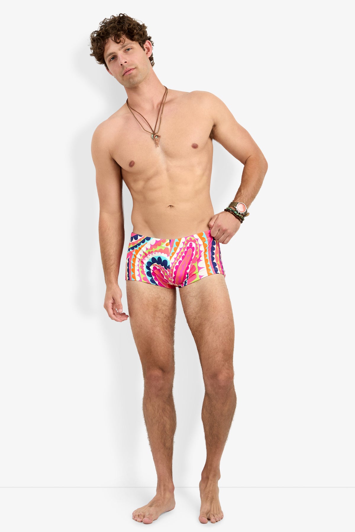 A man stands barefoot against a white background, wearing the Sorrento Swim Trunk featuring a colorful pink and blue pattern. Shirtless, he accessorizes with a bracelet and necklaces, exuding a relaxed expression and posture.