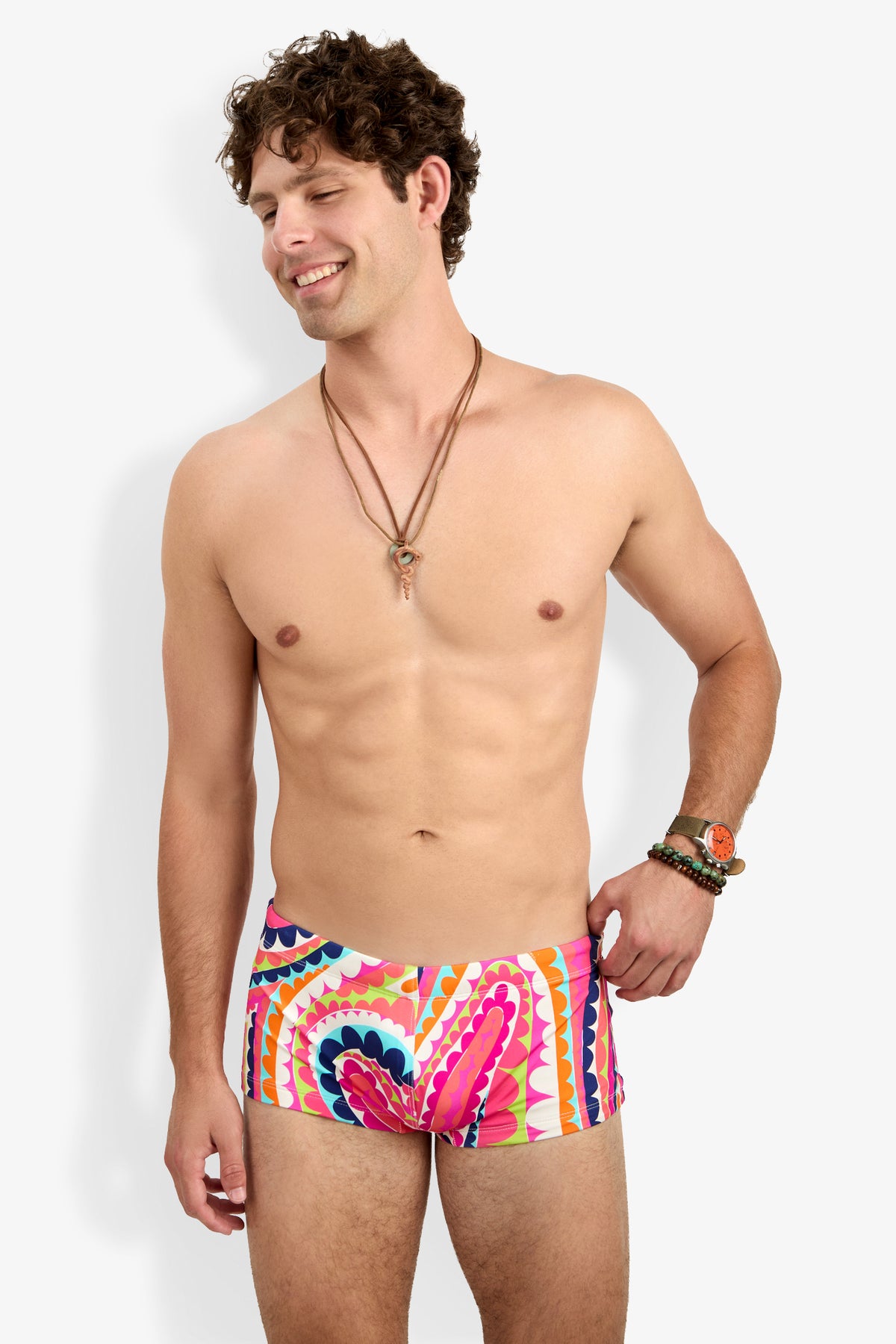 A smiling man with curly brown hair wears the Sorrento Swim Trunk, layered necklaces, and bracelets. He is shirtless and stands against a plain white background.