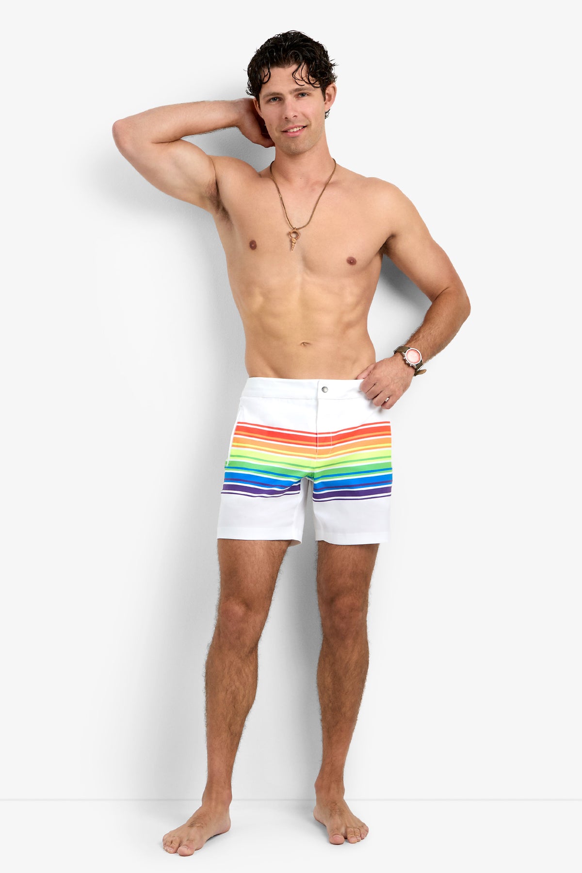 A man stands barefoot against a plain background, smiling and shirtless, wearing the Honolulu Board Short featuring white fabric with colorful horizontal rainbow stripes. One hand rests on his head while the other is by his side.