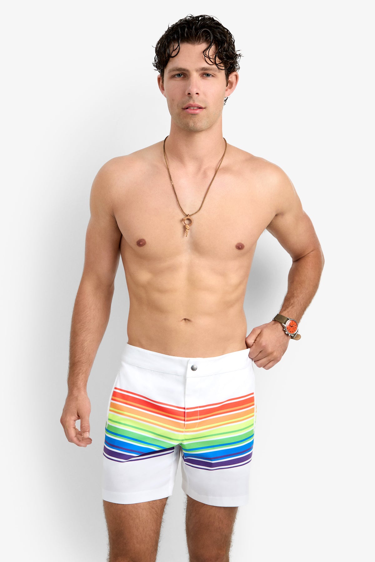 A shirtless man with short, wavy brown hair, a necklace, and an orange watch confidently models the Honolulu Board Short—white shorts with horizontal rainbow stripes—against a plain background.