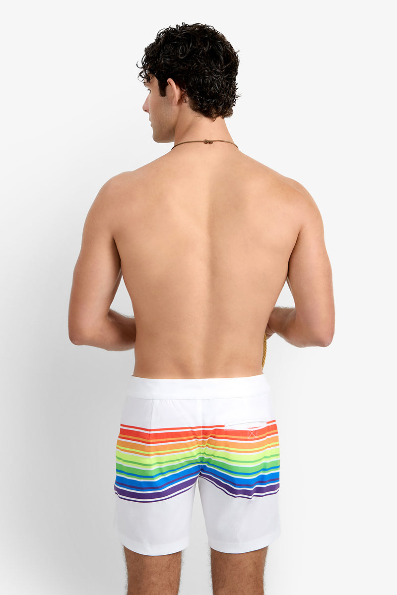 A shirtless man with short curly hair faces away, wearing the Honolulu Board Short—white swim trunks featuring horizontal rainbow stripes on the back—set against a plain white background.