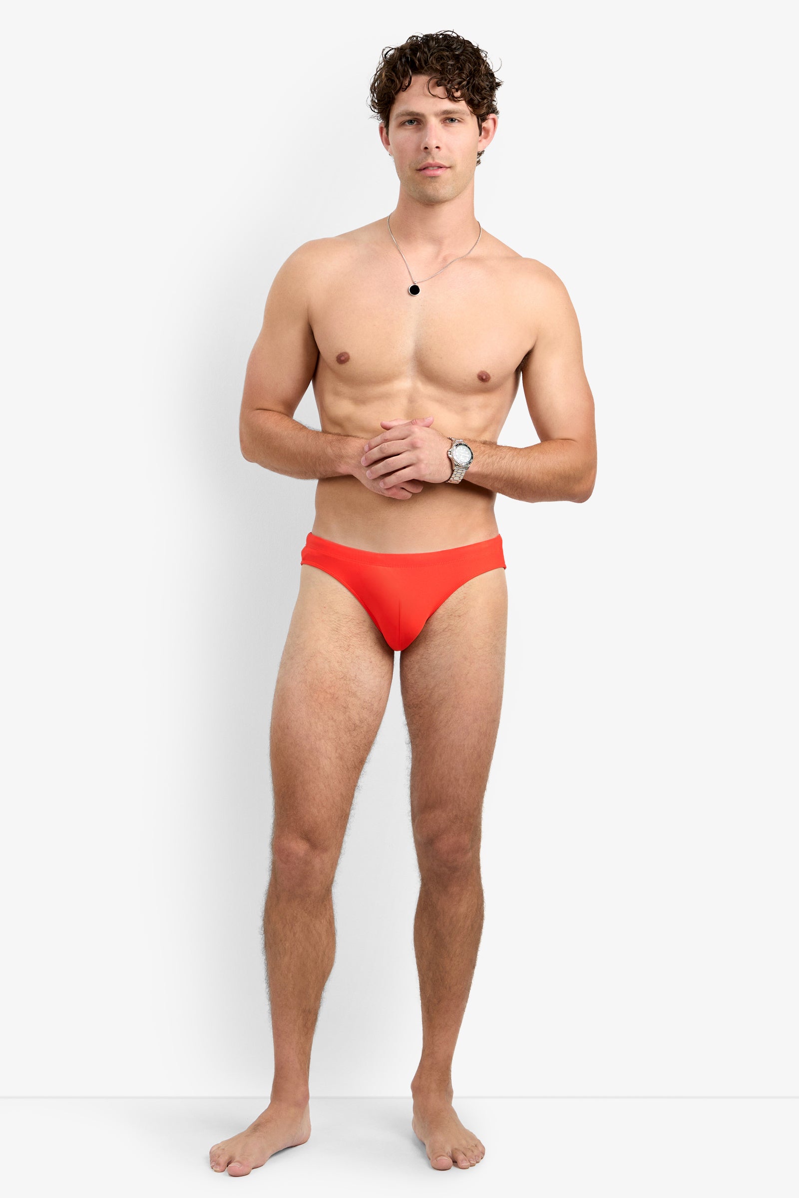 A young man with curly hair stands barefoot against a white background, wearing the Bondi Beach Swim Brief in bright red. He is shirtless, accessorized with a wristwatch and necklace, his hands clasped in front of him.