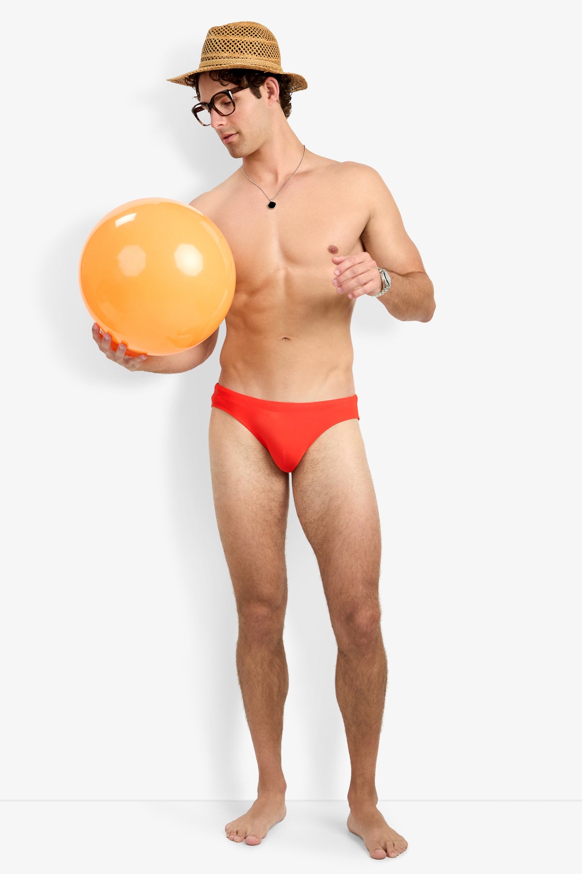 A man in Bondi Beach Swim Brief stands barefoot, wearing a straw hat, glasses, a necklace, and a watch, holding an orange beach ball and looking at it against a plain white background.