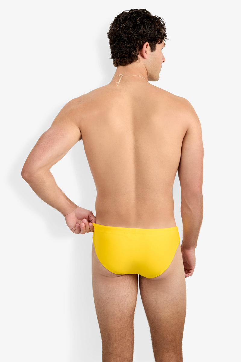A man with short, curly dark hair faces away, wearing the Bondi Swim Brief in bright yellow. His upper body is bare as he slightly pulls at the waistband against a plain white background.