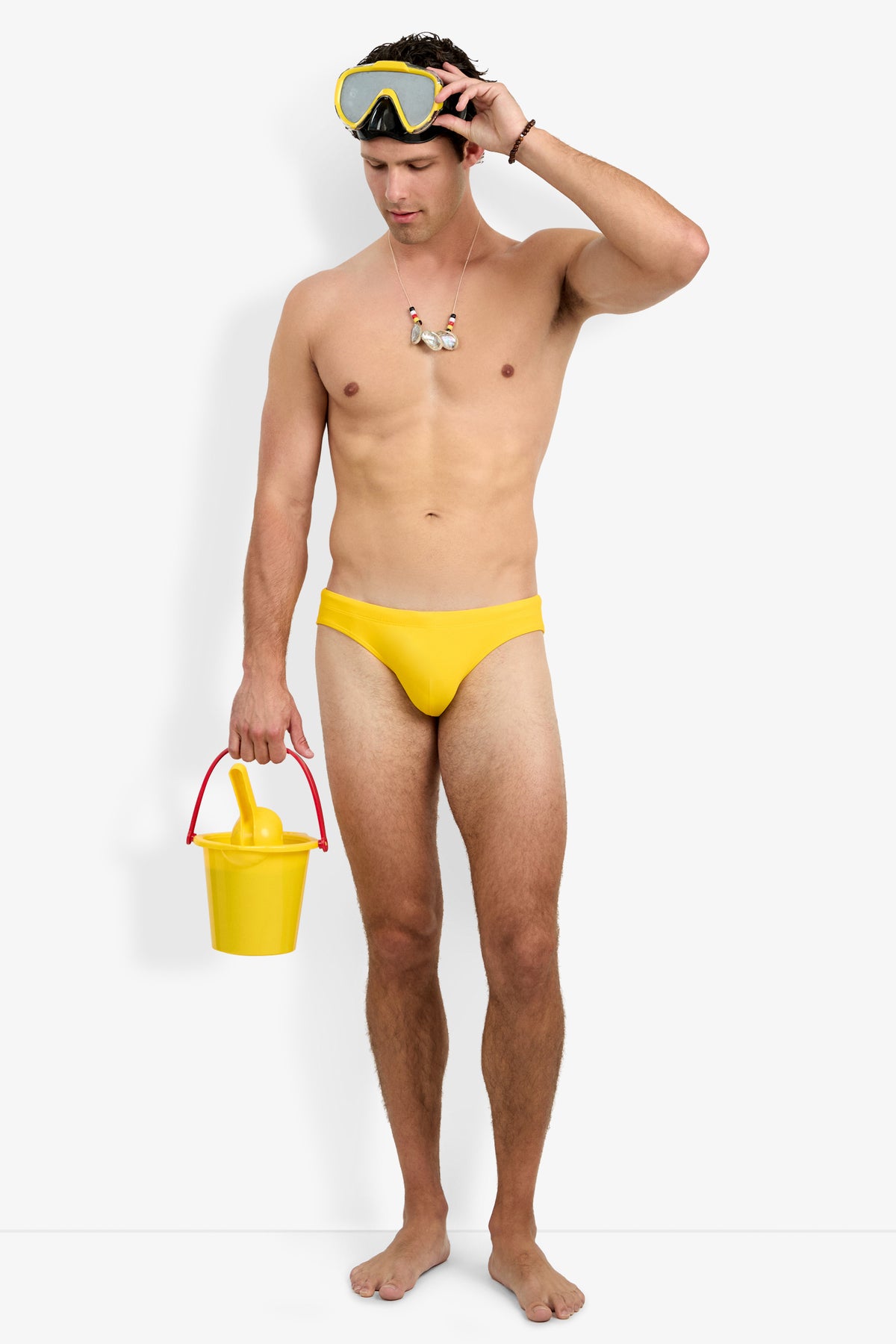 A shirtless man wears the yellow Bondi Swim Brief, a matching snorkel mask on his head, a seashell necklace, and holds a yellow bucket with a banana inside. He stands barefoot against a plain white background.